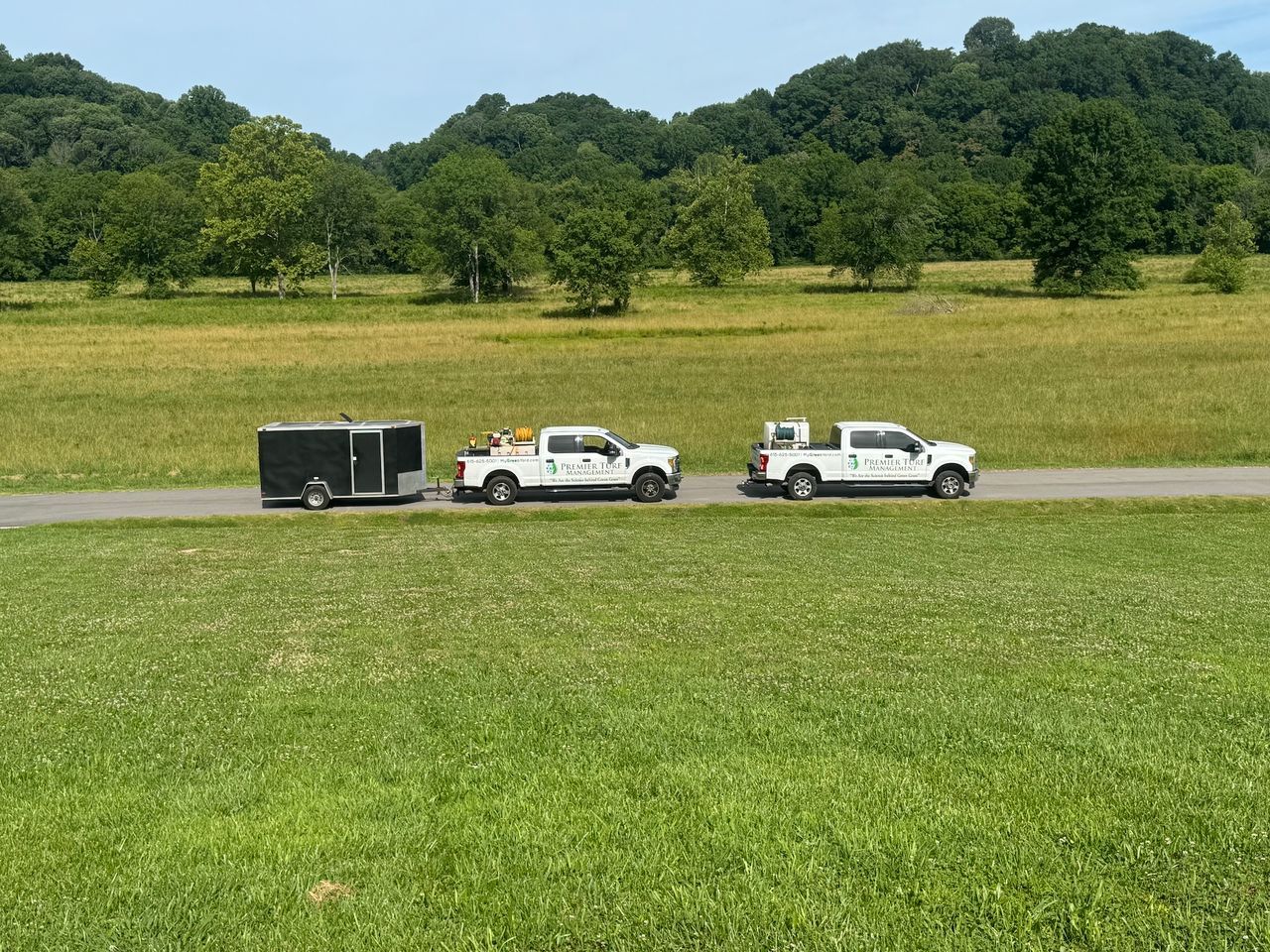 Two white pickup trucks towing a black trailer across a grassy field near trees
