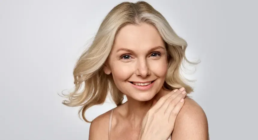 Blonde woman smiles, touching shoulder, against a light gray background.