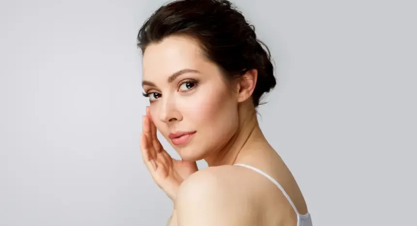 Woman with dark hair, touching cheek, looking over shoulder, neutral background.