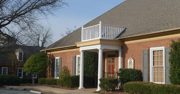 Brick building with a balcony, white columns, and shrubs under a sunny sky.