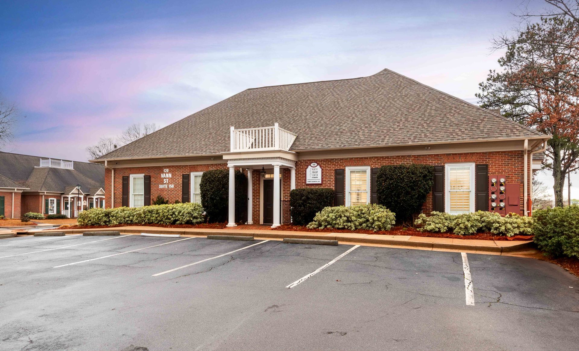 Brick building with a balcony, white columns, and shrubs under a sunny sky.