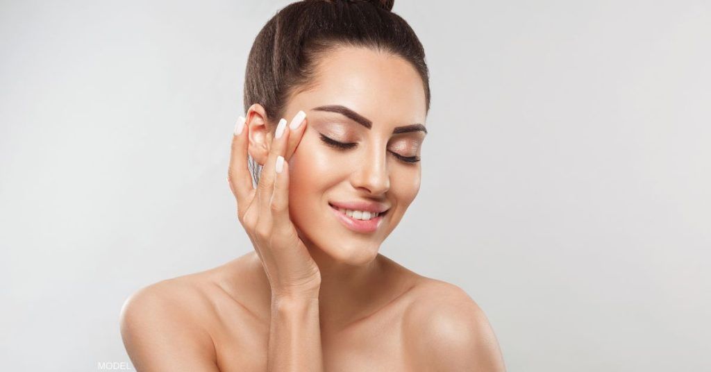 Woman touching her face, eyes closed, smiling, against a light gray background.