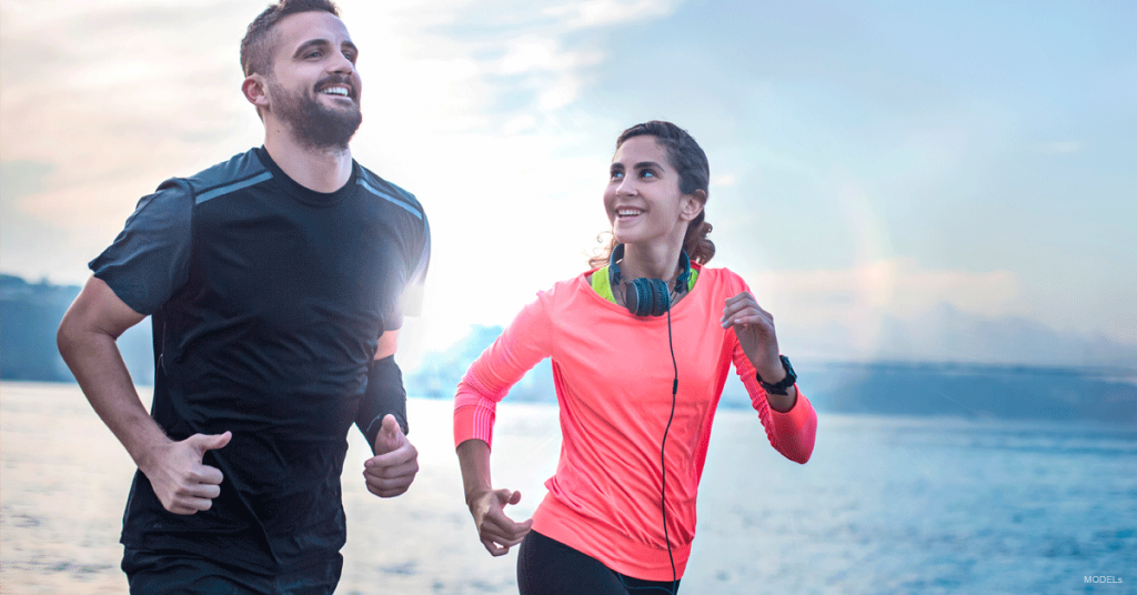 Man and woman running near water, both smiling. The woman wears pink, the man dark gray.