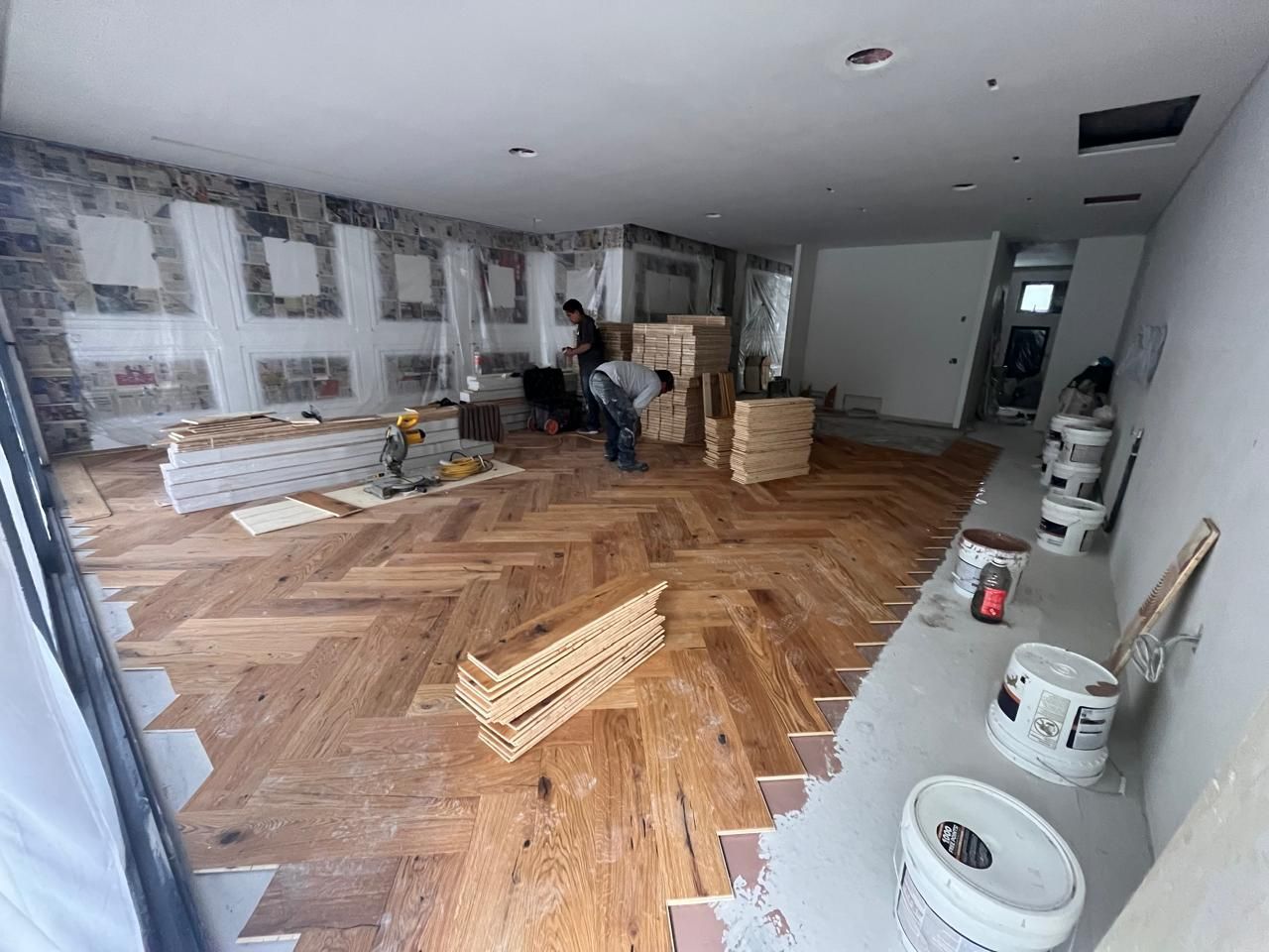 Habitación reformada con suelo de madera en espiga, materiales de construcción y trabajadores cerca de la pared del fondo.
