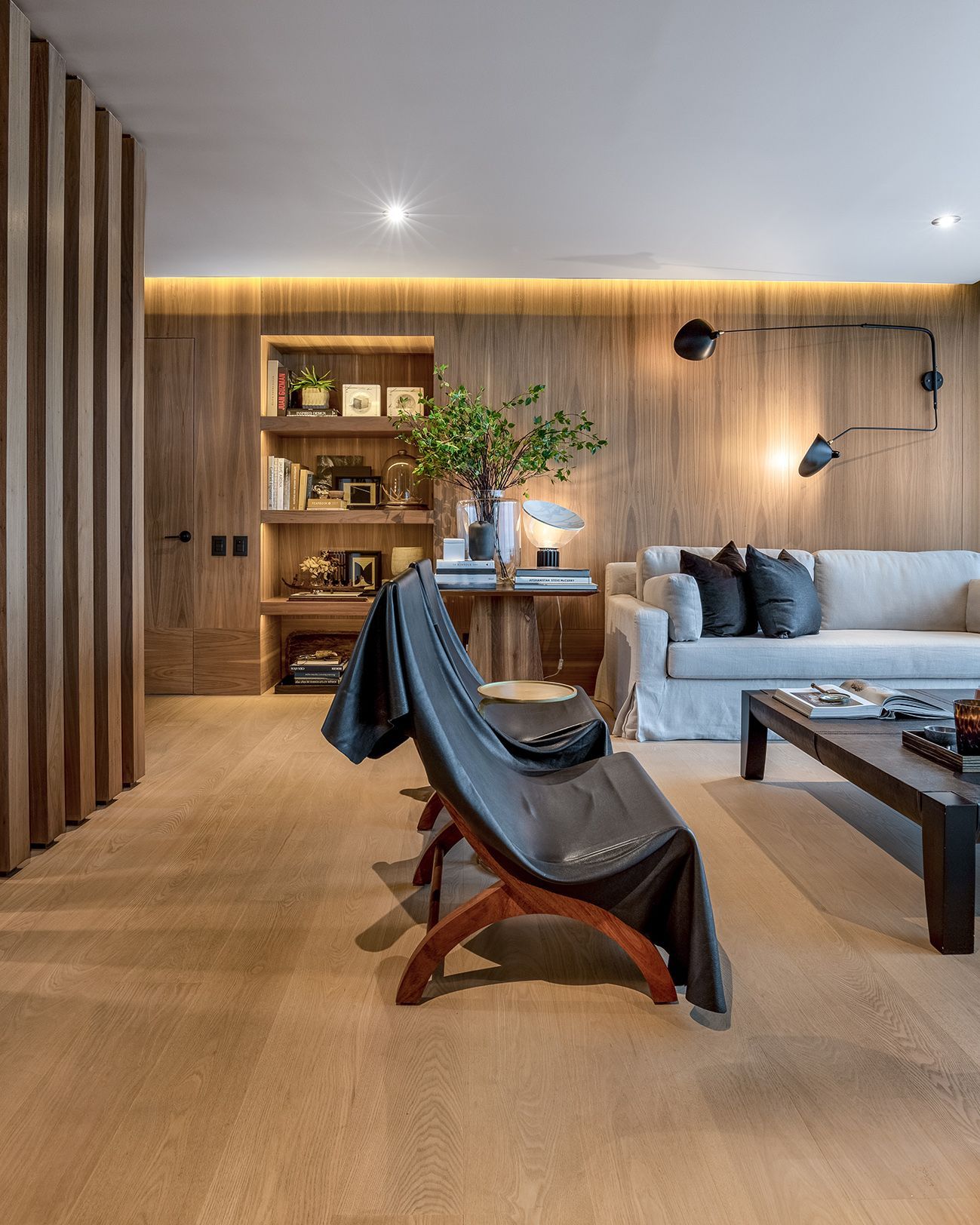 Sala de estar moderna con paredes de madera, sofá gris, sillón de cuero e iluminación empotrada cálida.