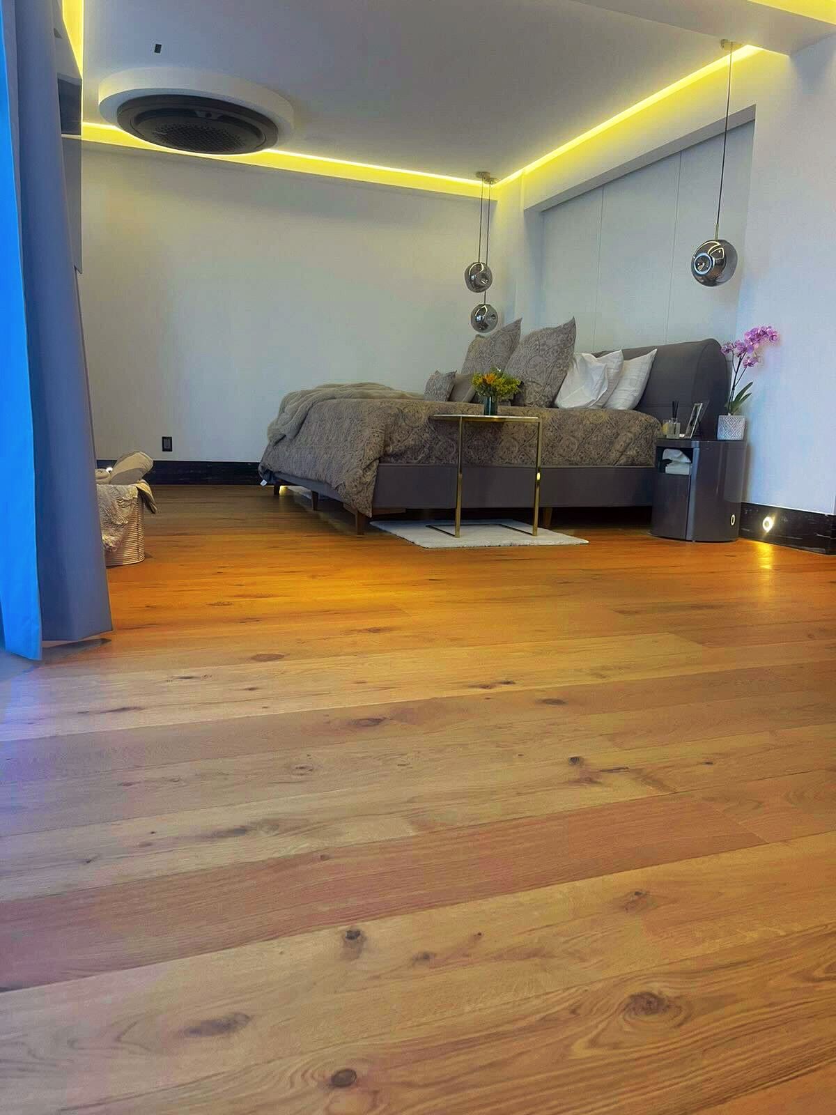 Dormitorio moderno con suelo de madera, cama gris, luces LED cálidas en el techo y una acogedora decoración minimalista.