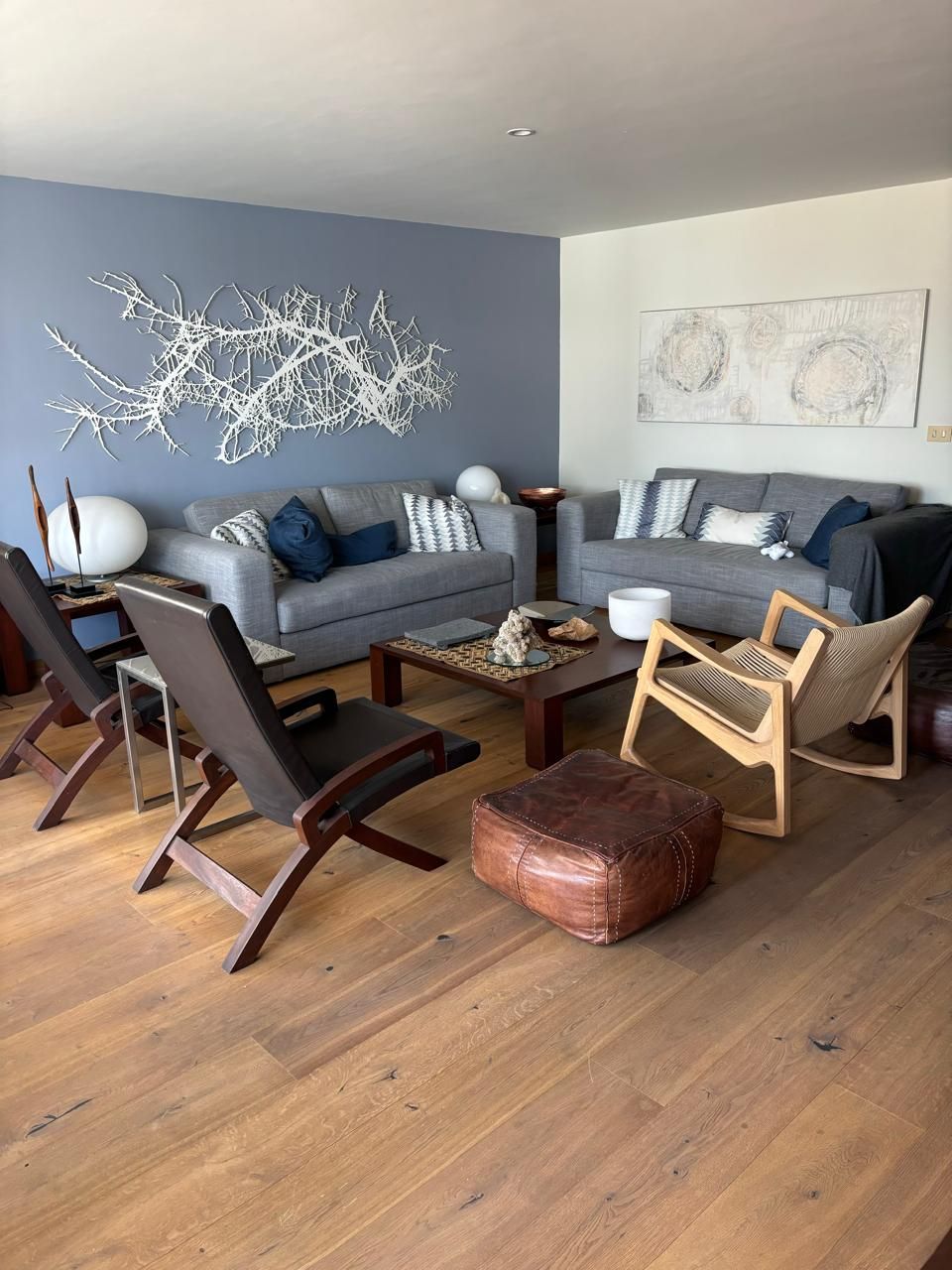 Sala de estar moderna con sofás grises, sillas de madera, mesa de centro y pared decorativa azul con cuadros.