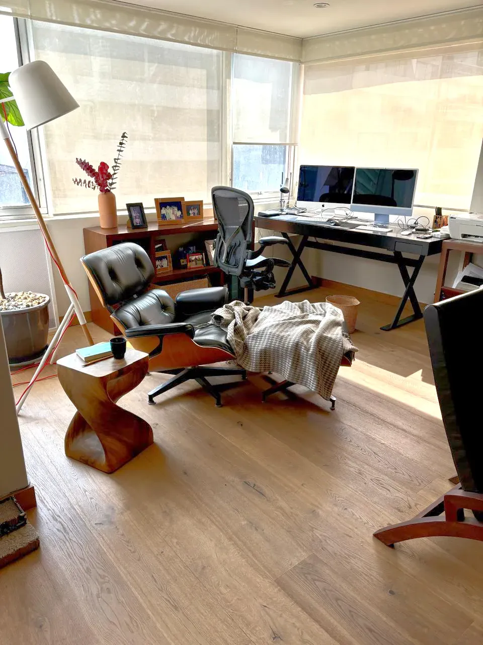 Luminoso despacho en casa con escritorio, monitores de ordenador, sillón, lámpara de pie y grandes ventanales.