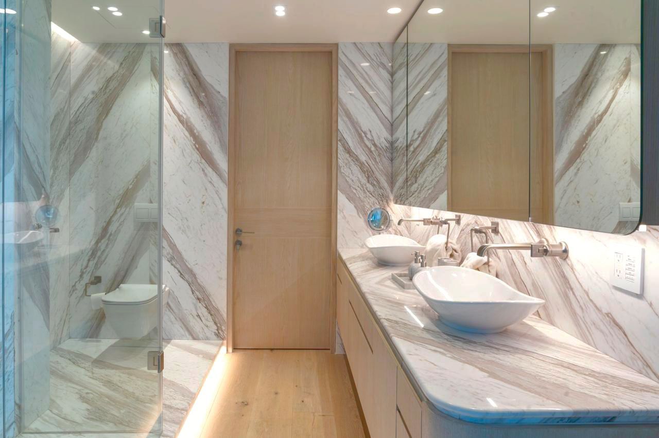 Baño moderno con paredes de mármol, detalles en madera, lavabo doble y mampara de ducha de cristal.