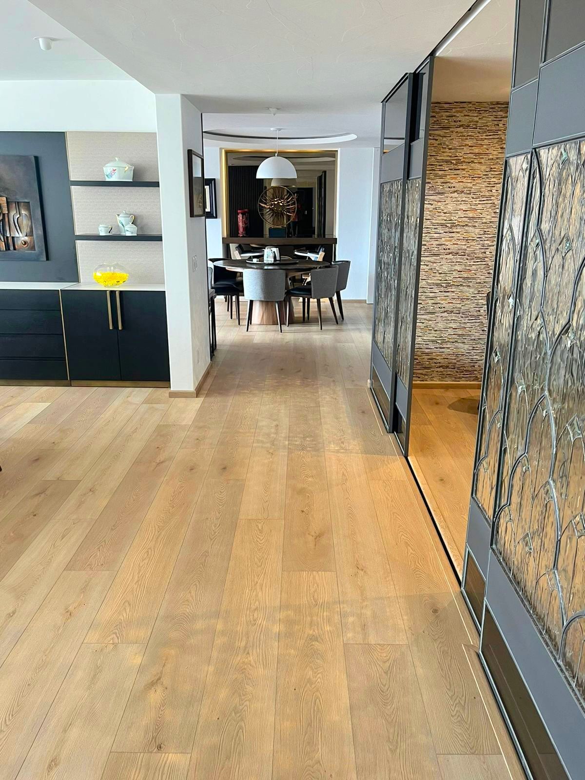 Pasillo moderno con suelos de madera clara, paredes grises, estanterías empotradas y zona de comedor al fondo.