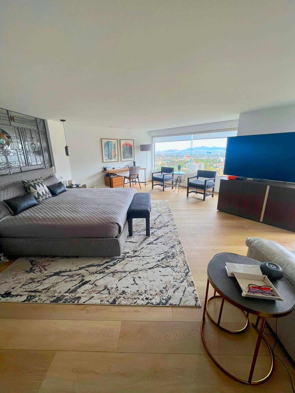 Sala de estar moderna con sofá modular gris, alfombra estampada, mesa de centro redonda y vista al mar a través de grandes ventanales.