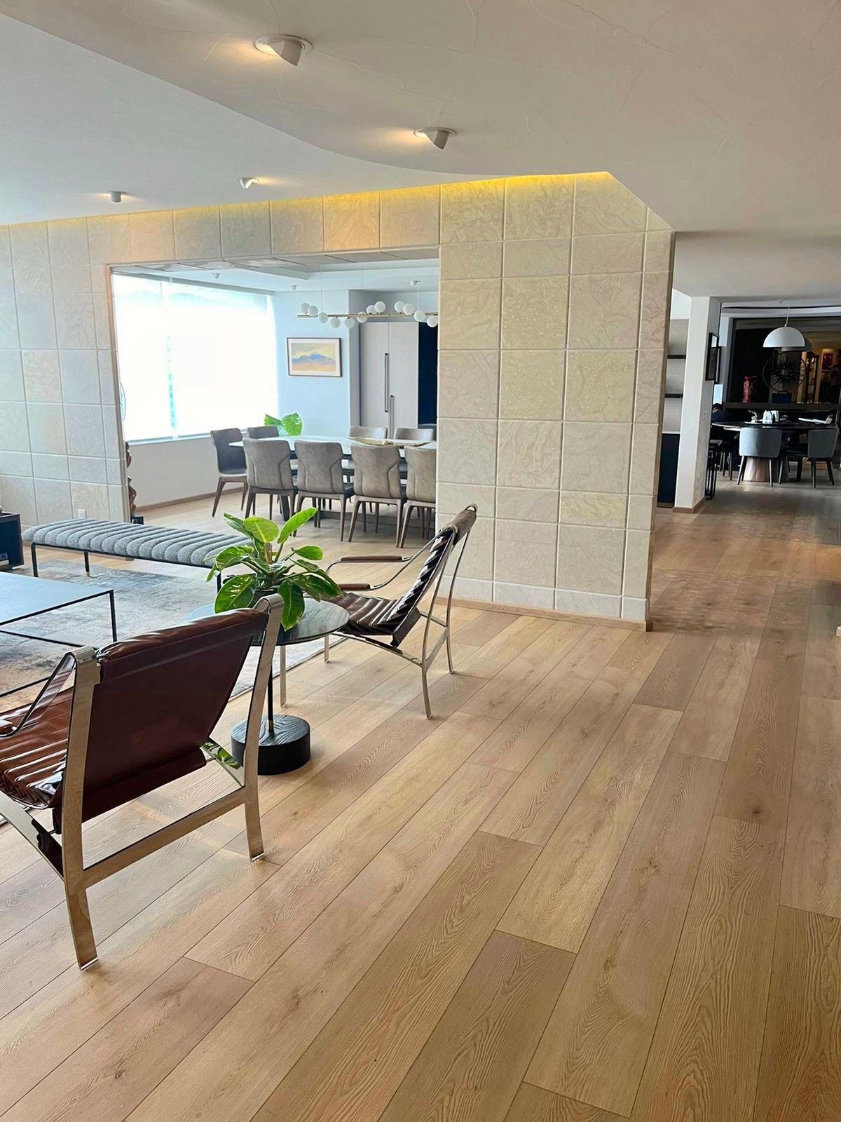 Salón moderno con suelos de madera, pared de piedra blanca, zona de comedor y asientos junto a una ventana luminosa.
