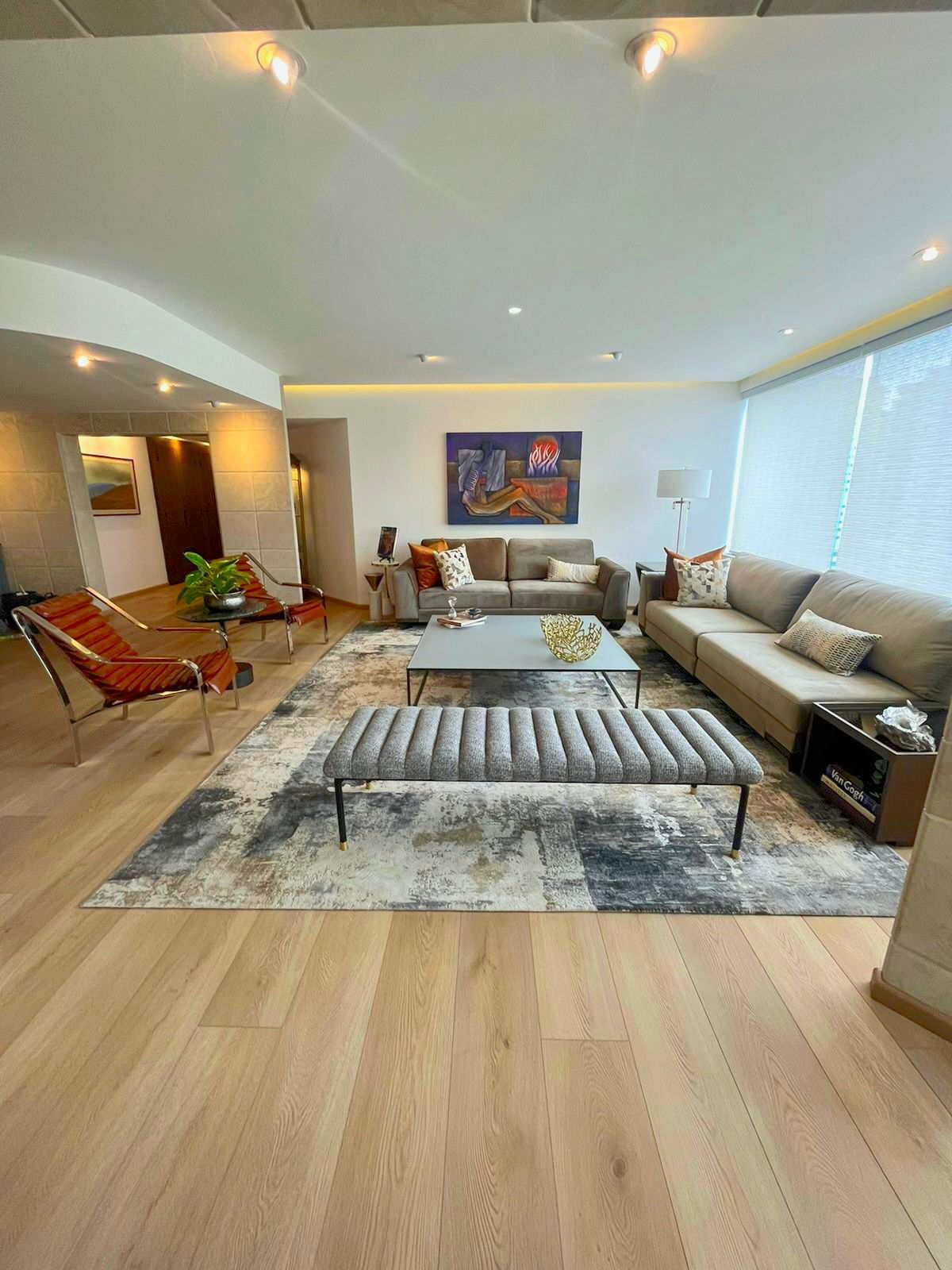 Sala de estar moderna con sofá modular, otomana a rayas, alfombra, suelo de madera y grandes ventanales.
