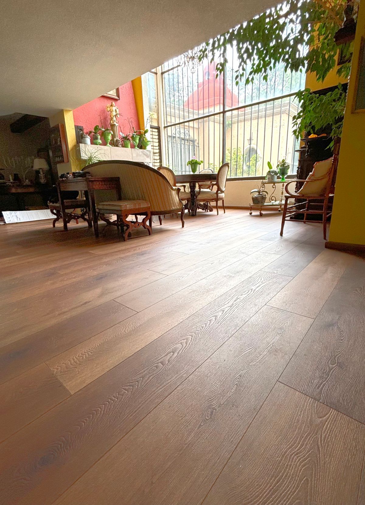 Patio cubierto con suelo de madera, sillas y mesas de mimbre, y vegetación junto a una valla metálica.