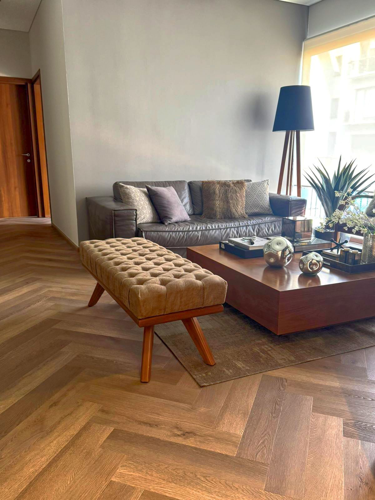 Sala de estar moderna con sofá gris, mesa de centro de madera, lámpara de pie azul y banco capitoné sobre suelo de madera en espiga.