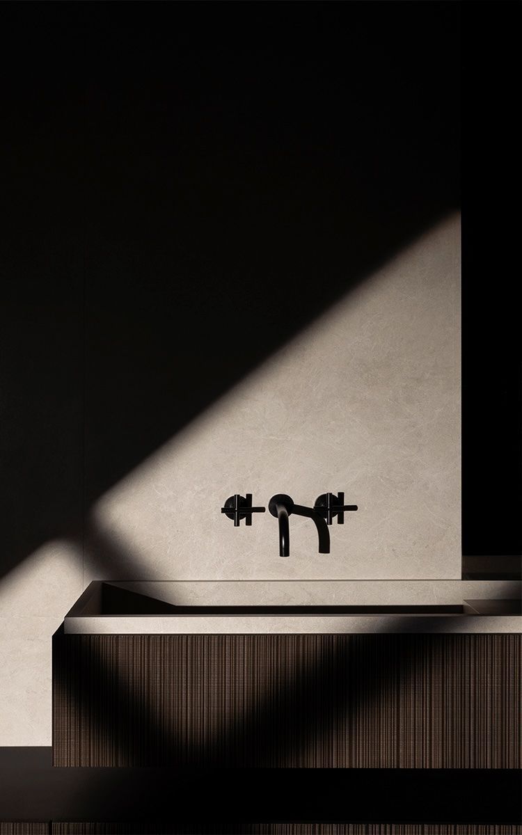 Lavabo minimalista con grifo negro sobre una encimera clara, iluminado por un rayo de sol diagonal sobre una pared oscura.