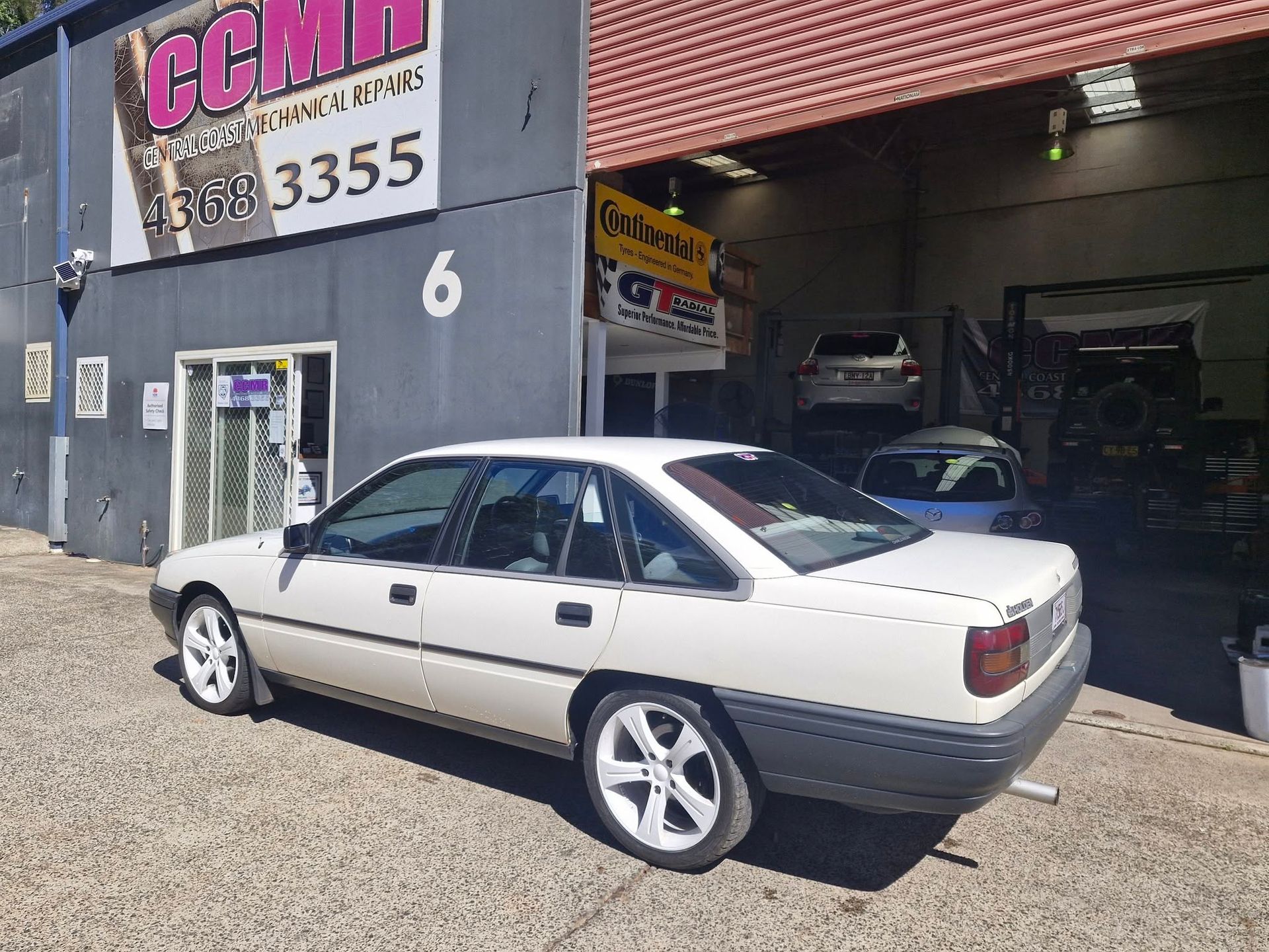 White coupe parked outside a garage with a pink CCM sign and open workshop bay