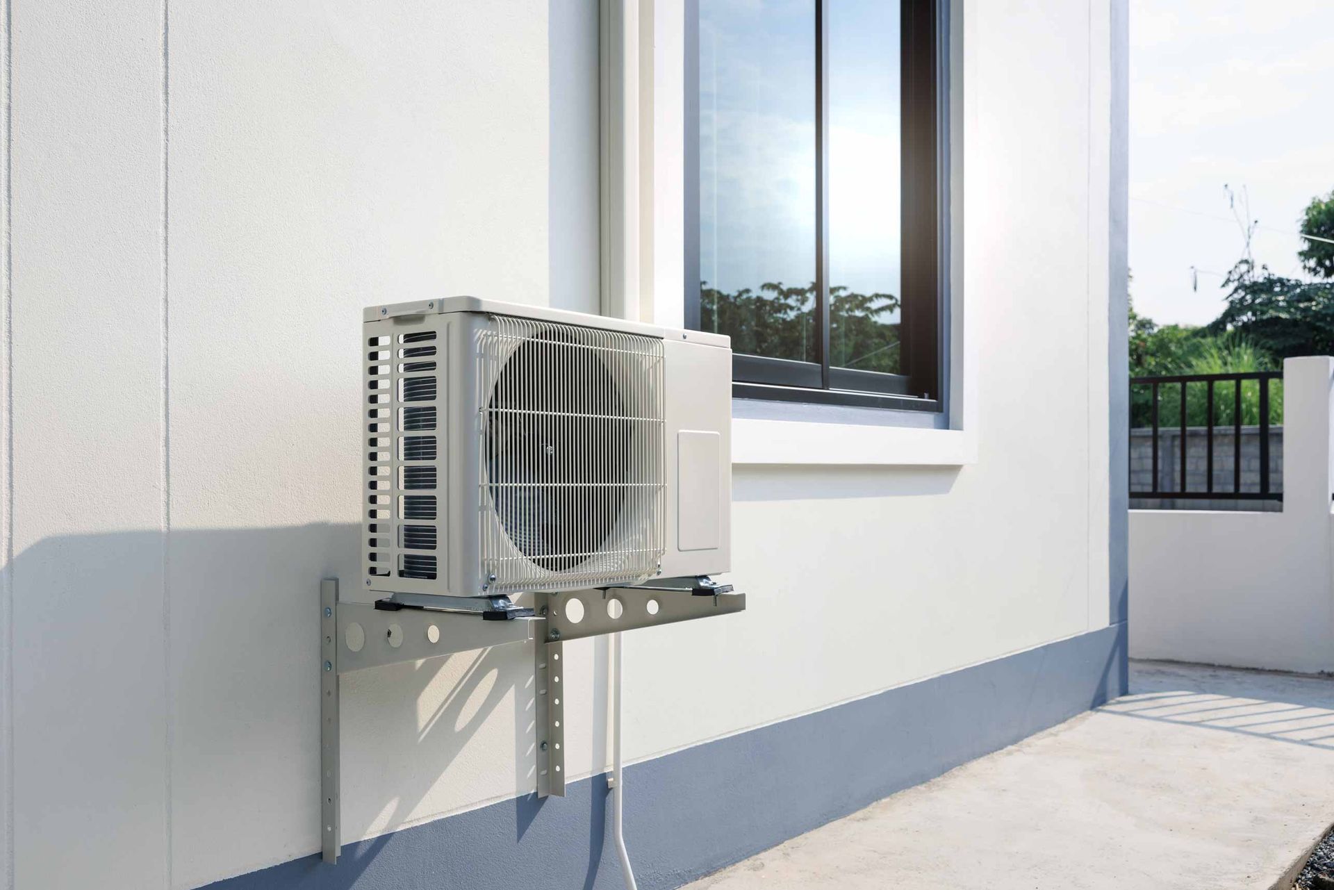 Outdoor air-conditioning unit mounted on a white wall beside a window and walkway