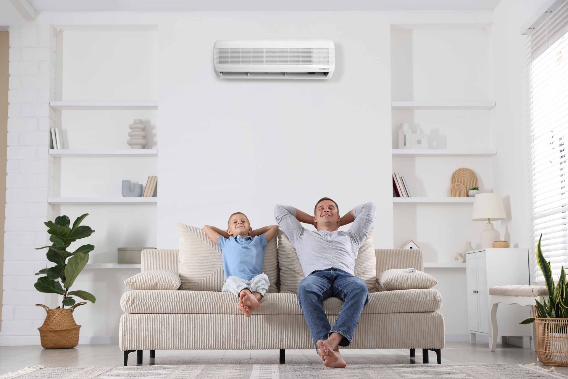Two people relaxing on a beige couch in a bright living room with white shelves and a wall-mounted air conditioner