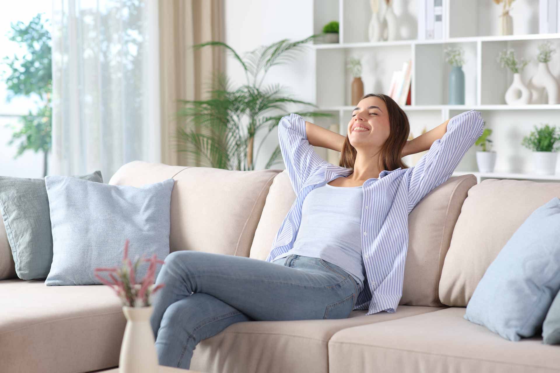 Woman relaxing on a beige sofa in a bright living room, hands behind head, smiling.