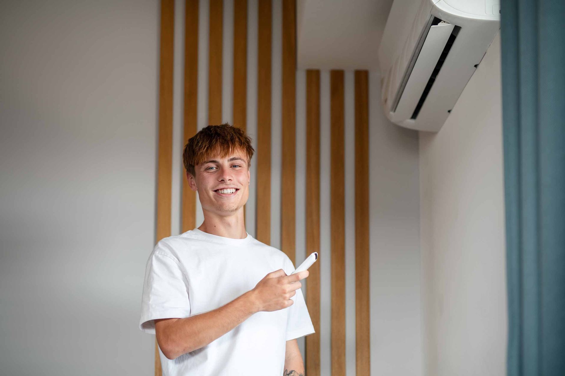 Person smiling and pointing at a wall-mounted air conditioner in a modern room with wood slats and blue curtains