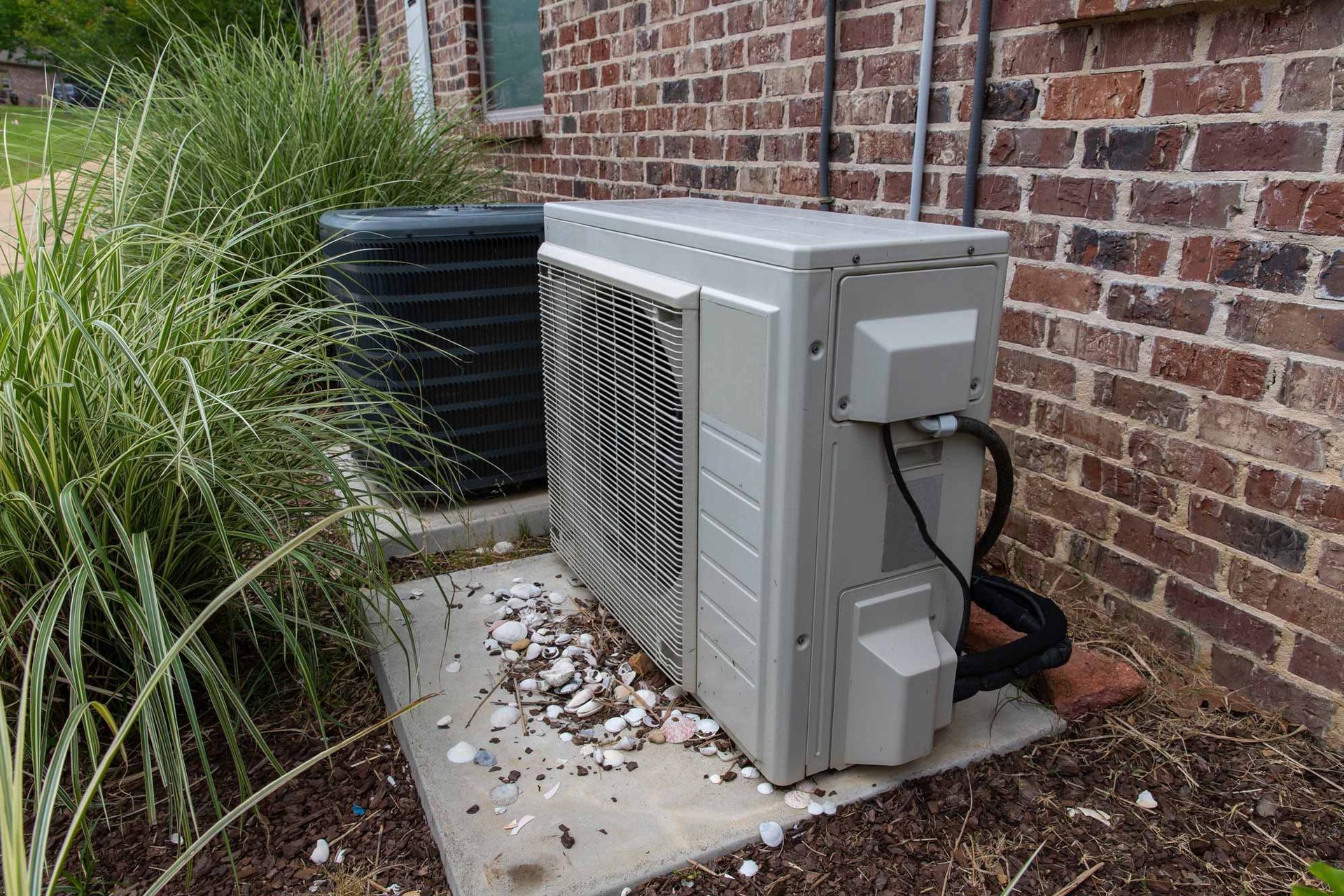 Outdoor air conditioning unit beside a brick house wall, with a black rain barrel and plants nearby