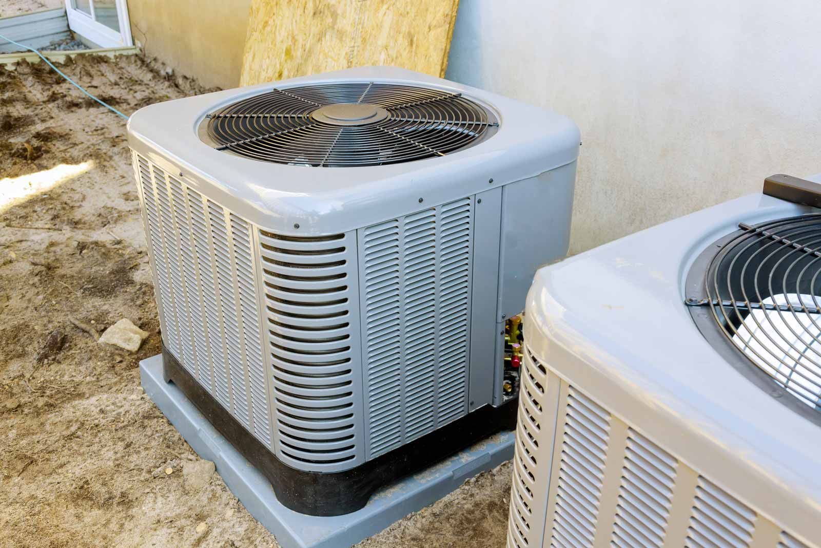 Two outdoor air conditioner units on concrete pads beside a beige wall and sandy ground