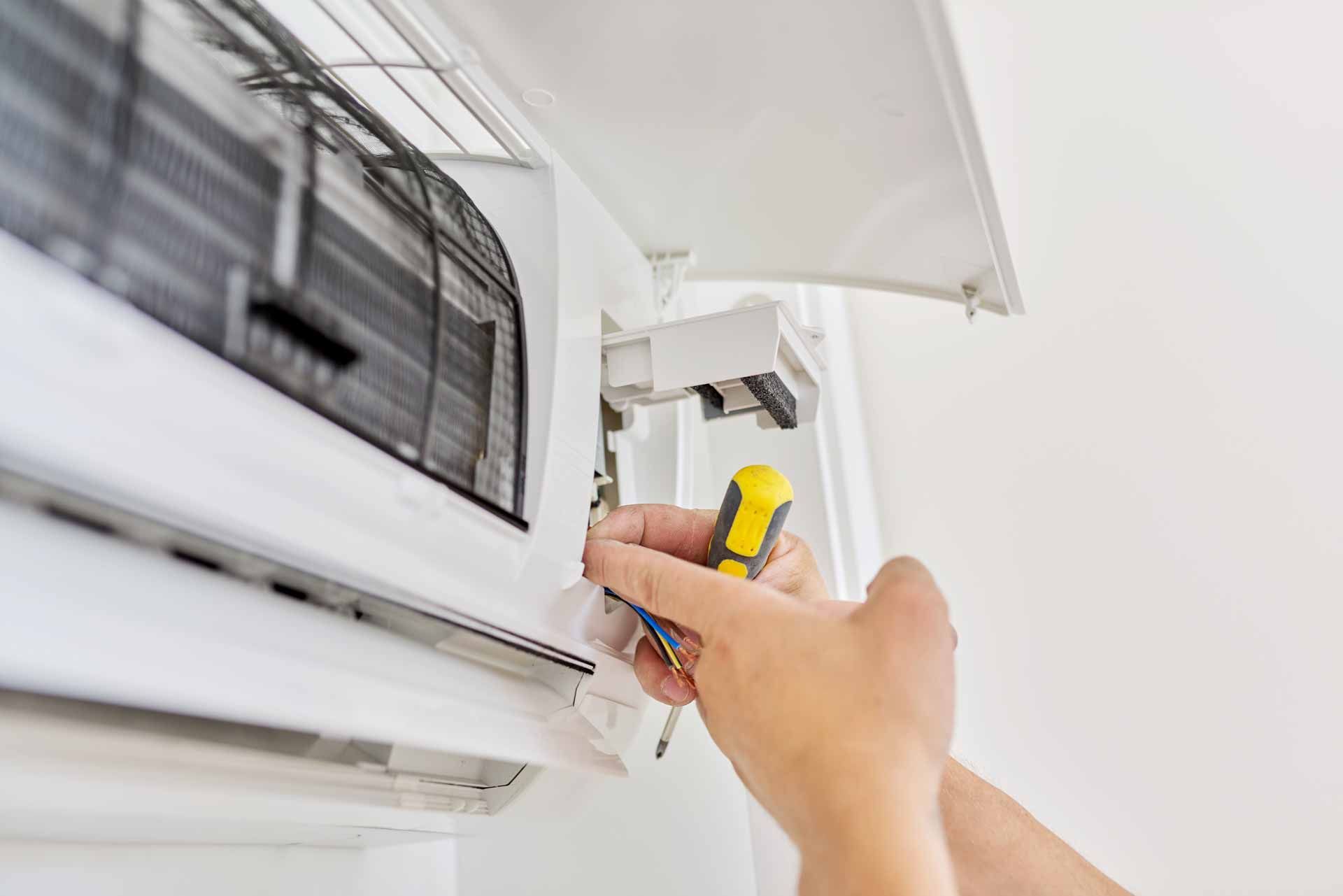 Hand opening an air conditioner panel with a yellow screwdriver under a white awning