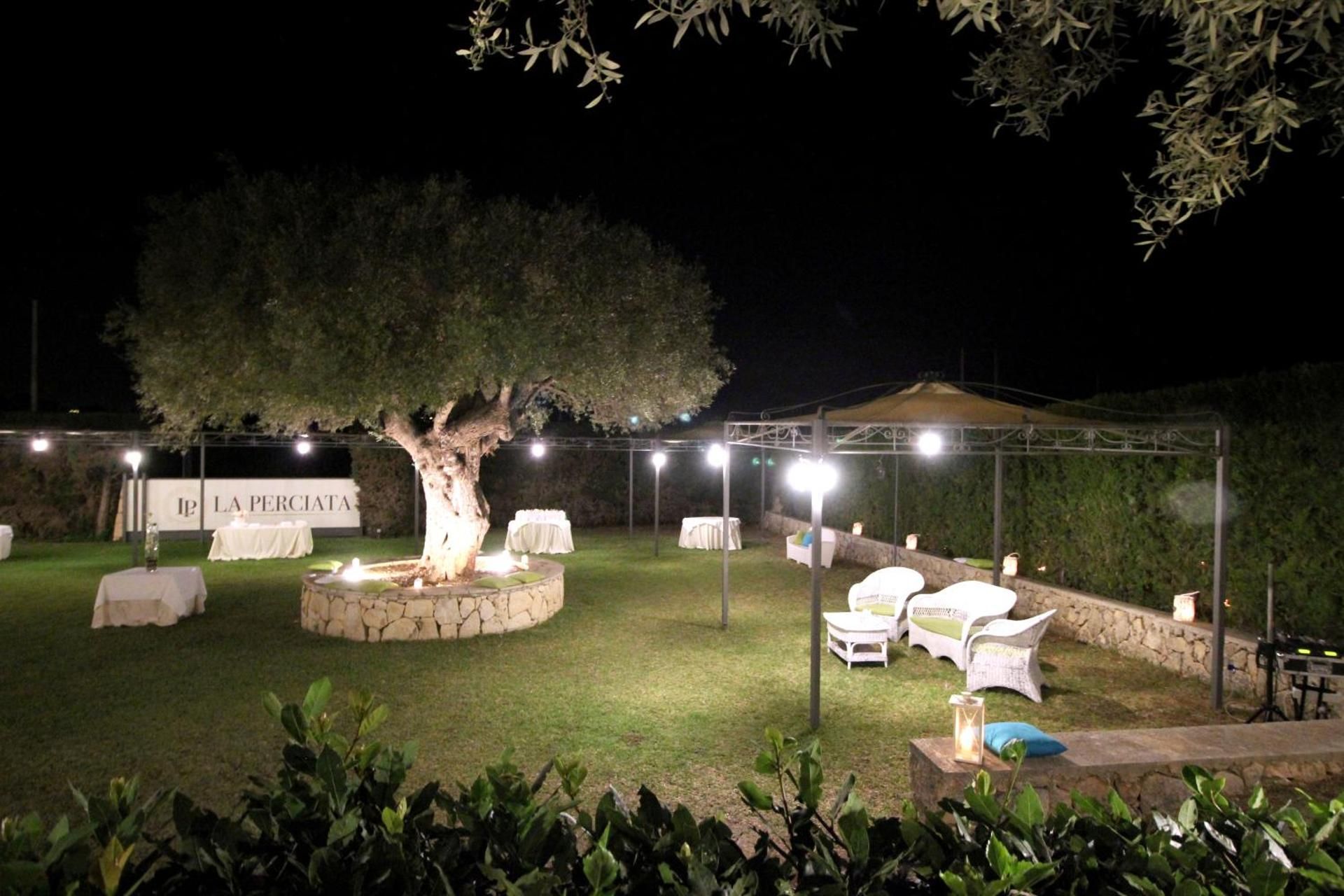 Patio notturno in giardino con albero illuminato, prato e sedie da esterno bianche sotto luci decorative.