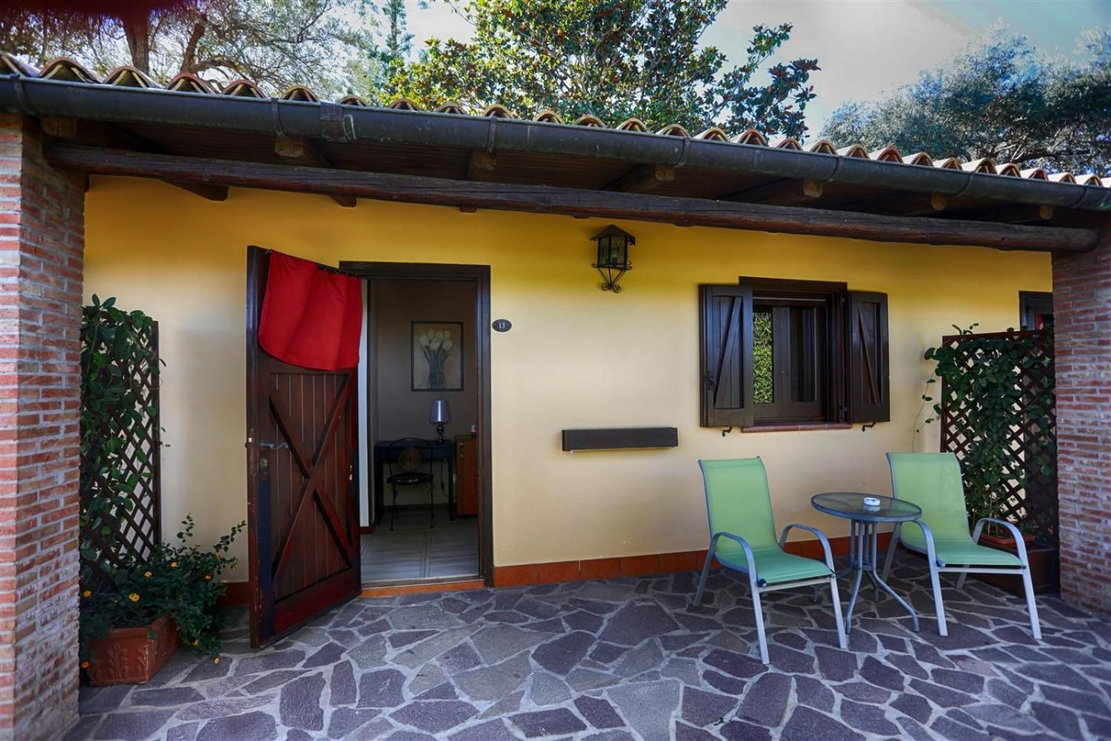 Veranda di un piccolo cottage giallo con porta aperta, sedie verdi e patio in pietra.