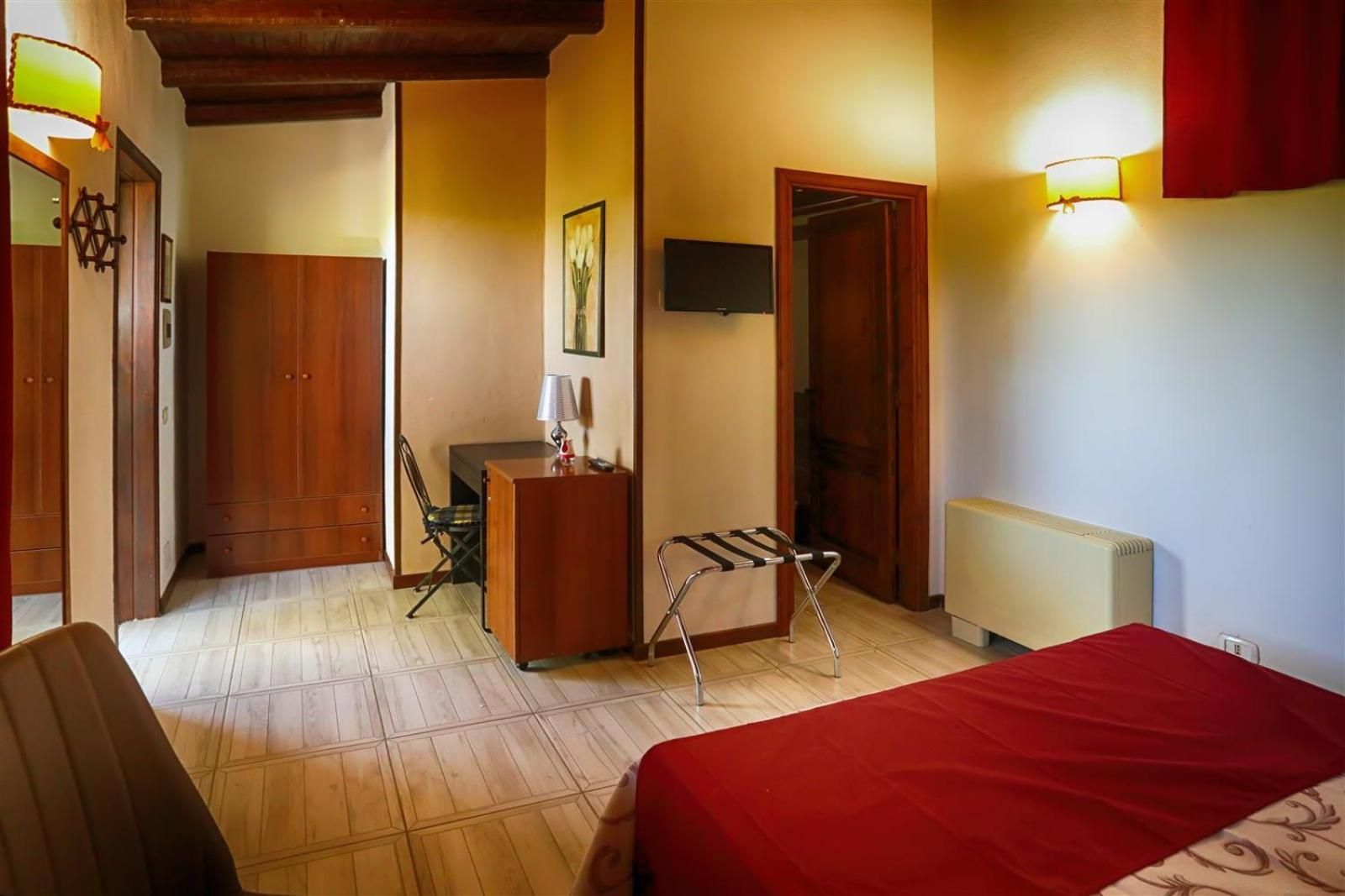 Camera da letto accogliente con copriletto rosso, pavimento in legno, armadio, sedia e porta in una stanza simile a quella di un hotel.