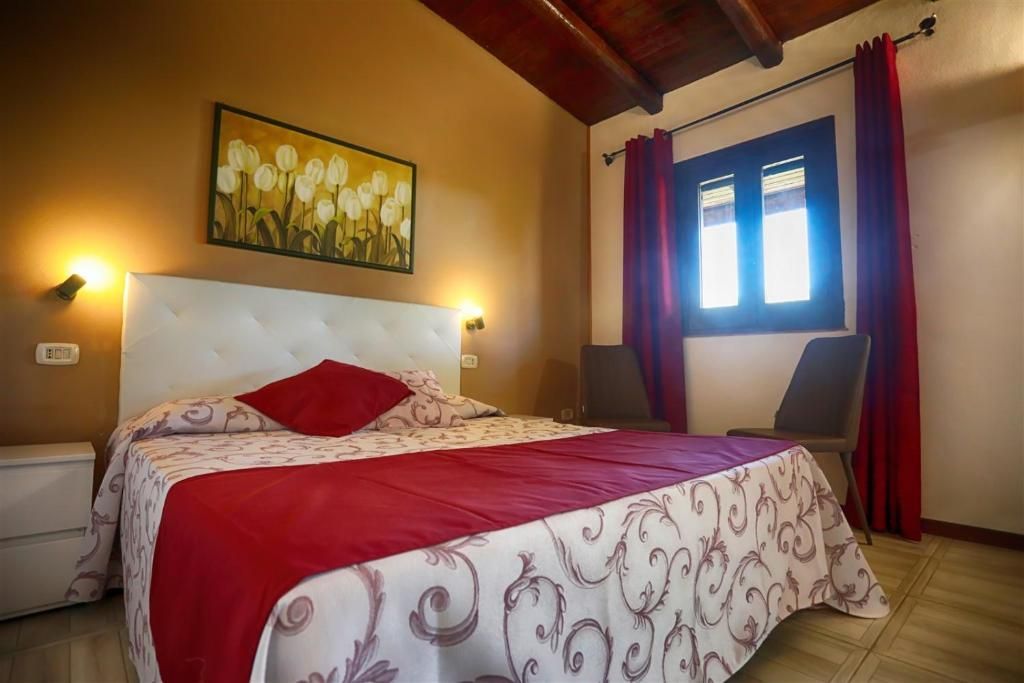 Accogliente camera da letto con copriletto rosso, travi a vista sul soffitto, lampade da parete e una finestra soleggiata.