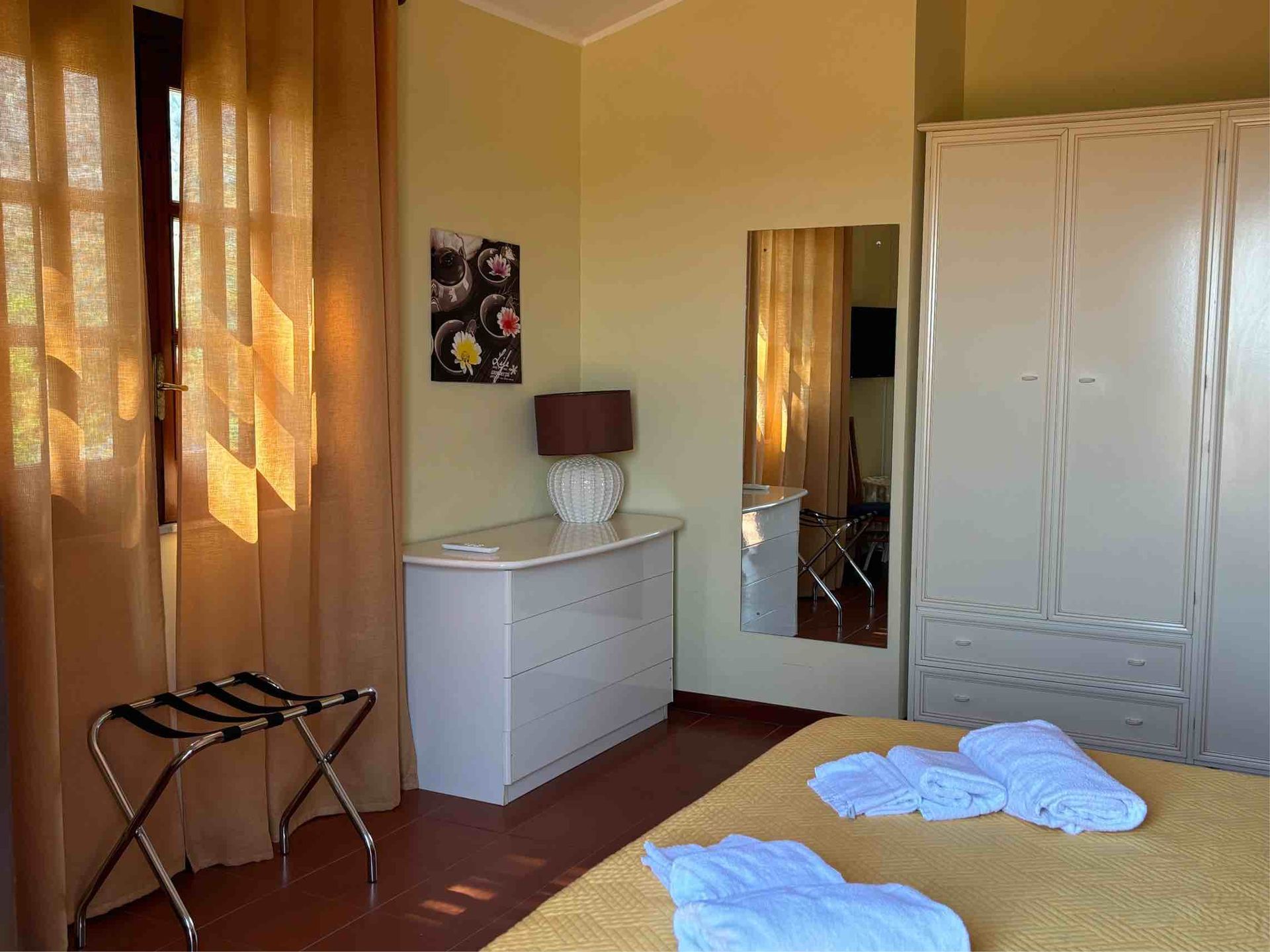 Camera da letto soleggiata con tende arancioni, comò bianco, specchio, armadio e copriletto stropicciato sul pavimento.