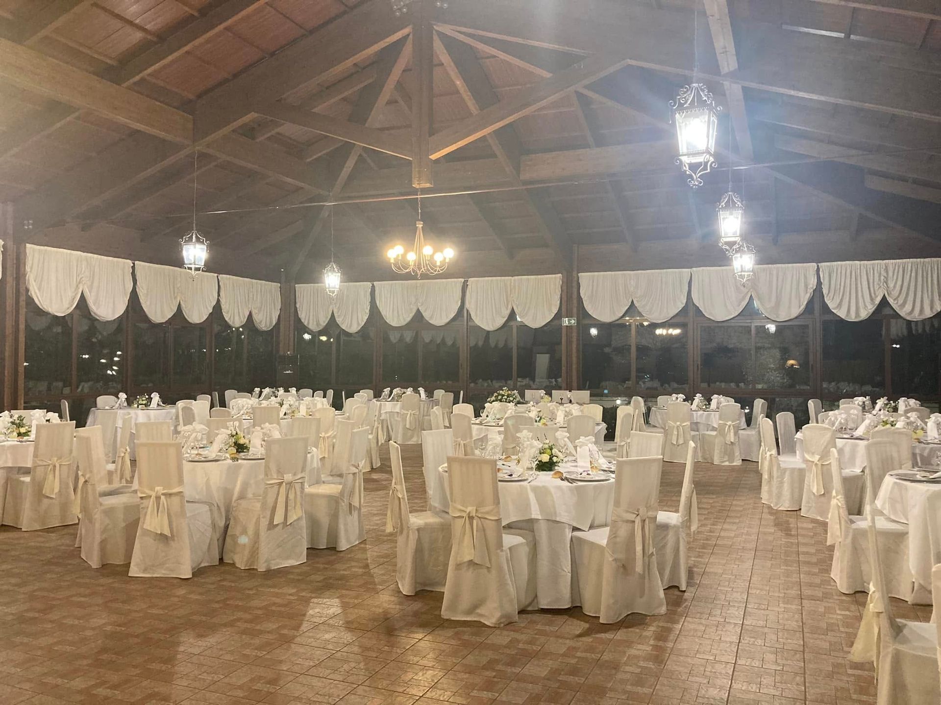 Sala da banchetto vuota con tavoli rotondi e sedie drappeggiati di bianco, illuminata da lampade a sospensione e con soffitto a volta in legno.