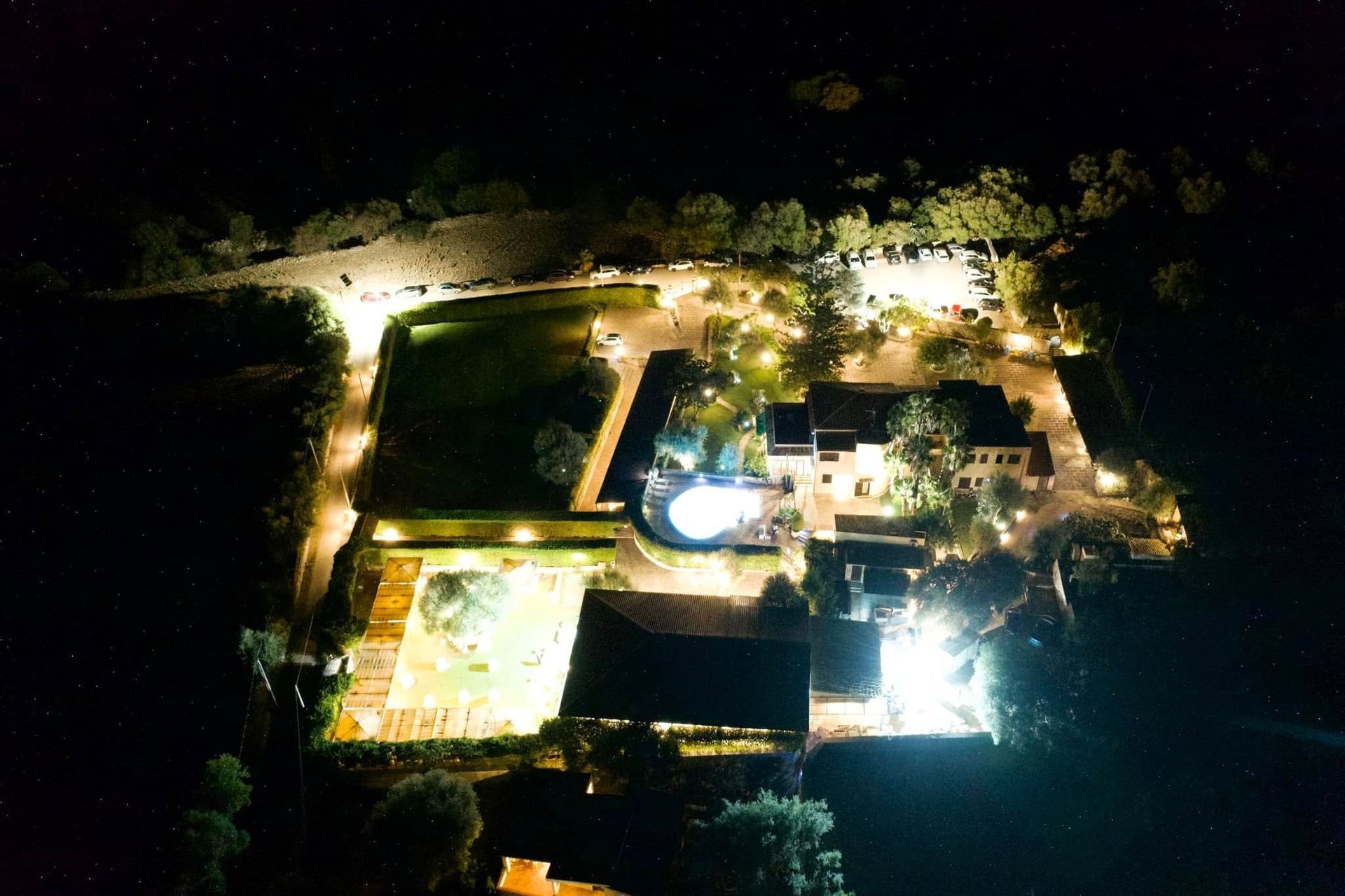 Veduta aerea notturna di una casa e del suo giardino illuminati a giorno, circondati da alberi scuri.