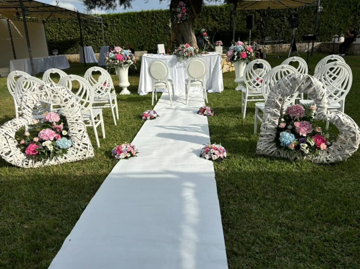 Una navata nuziale bianca con composizioni floreali e sedie bianche decorate su un prato erboso.