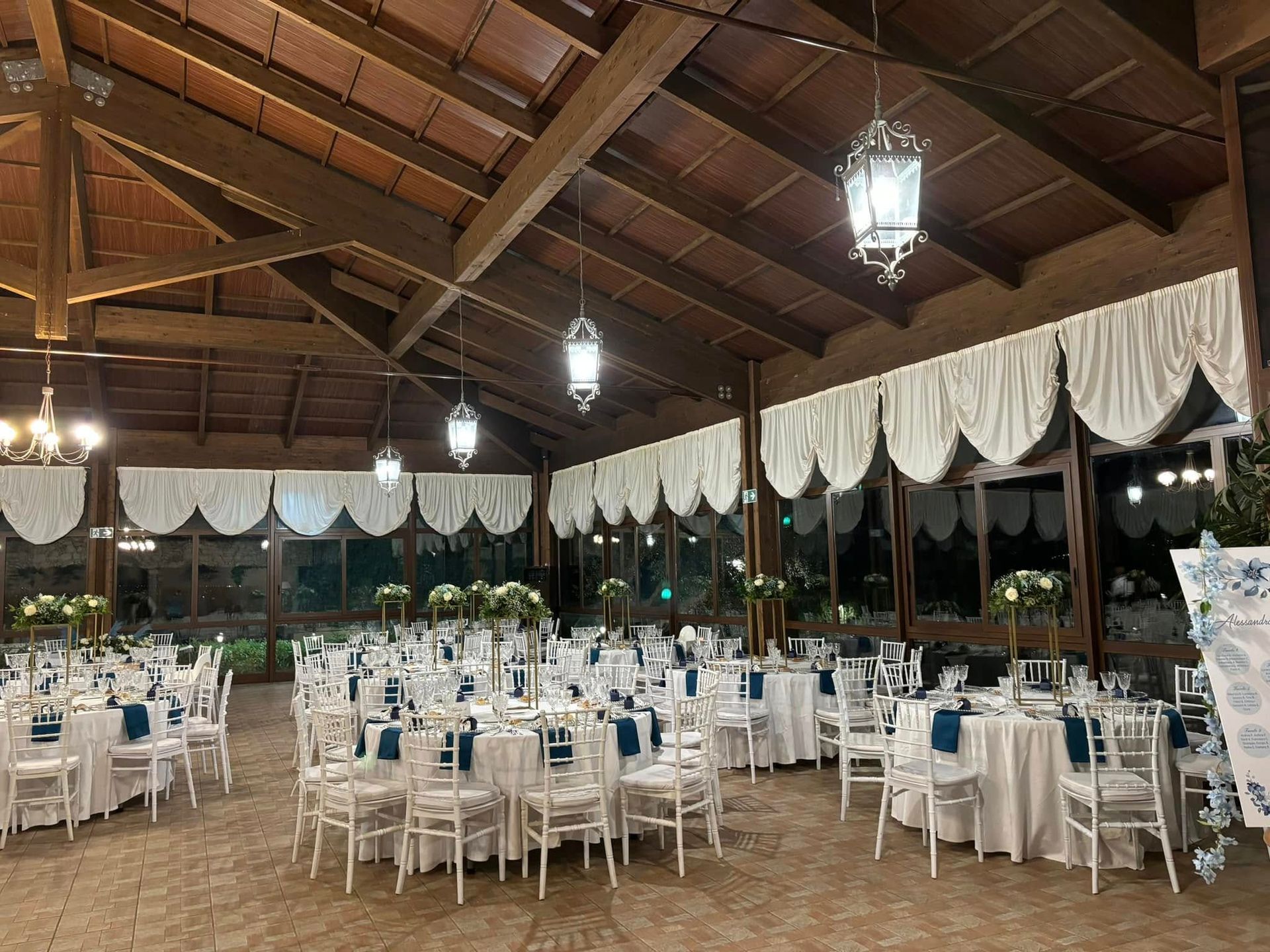 Sala da banchetto decorata con tavoli rotondi, sedie bianche e lanterne sospese sotto un soffitto di legno.