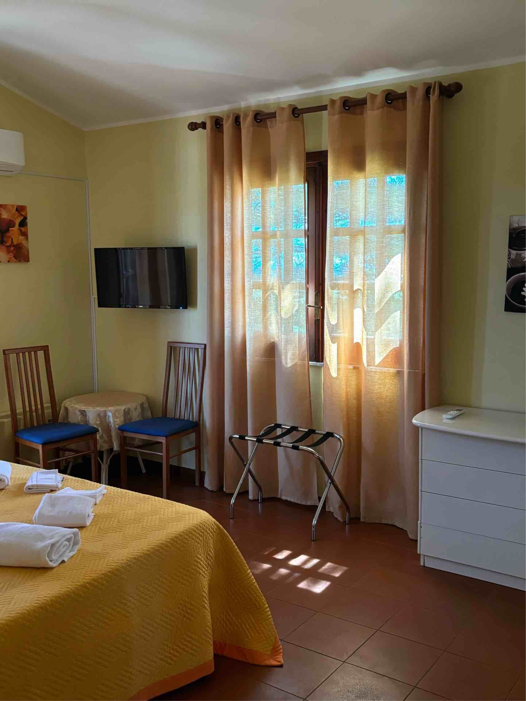 Camera da letto soleggiata con letto, sedie, TV a parete e cassettiera accanto a una finestra con tenda.