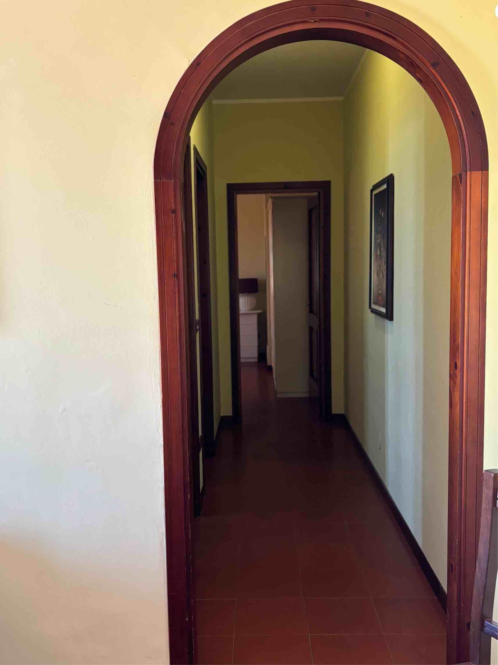 Stretto corridoio con pareti gialle, pavimento rosso e una porta di legno ad arco in primo piano.