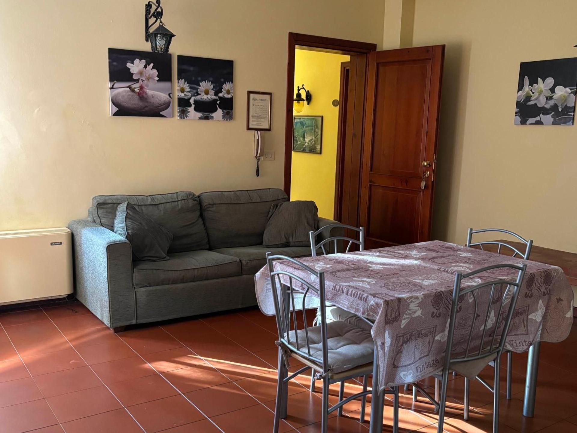 Piccolo soggiorno con divano grigio, tavolo da pranzo, quadri alle pareti e una porta di legno aperta.