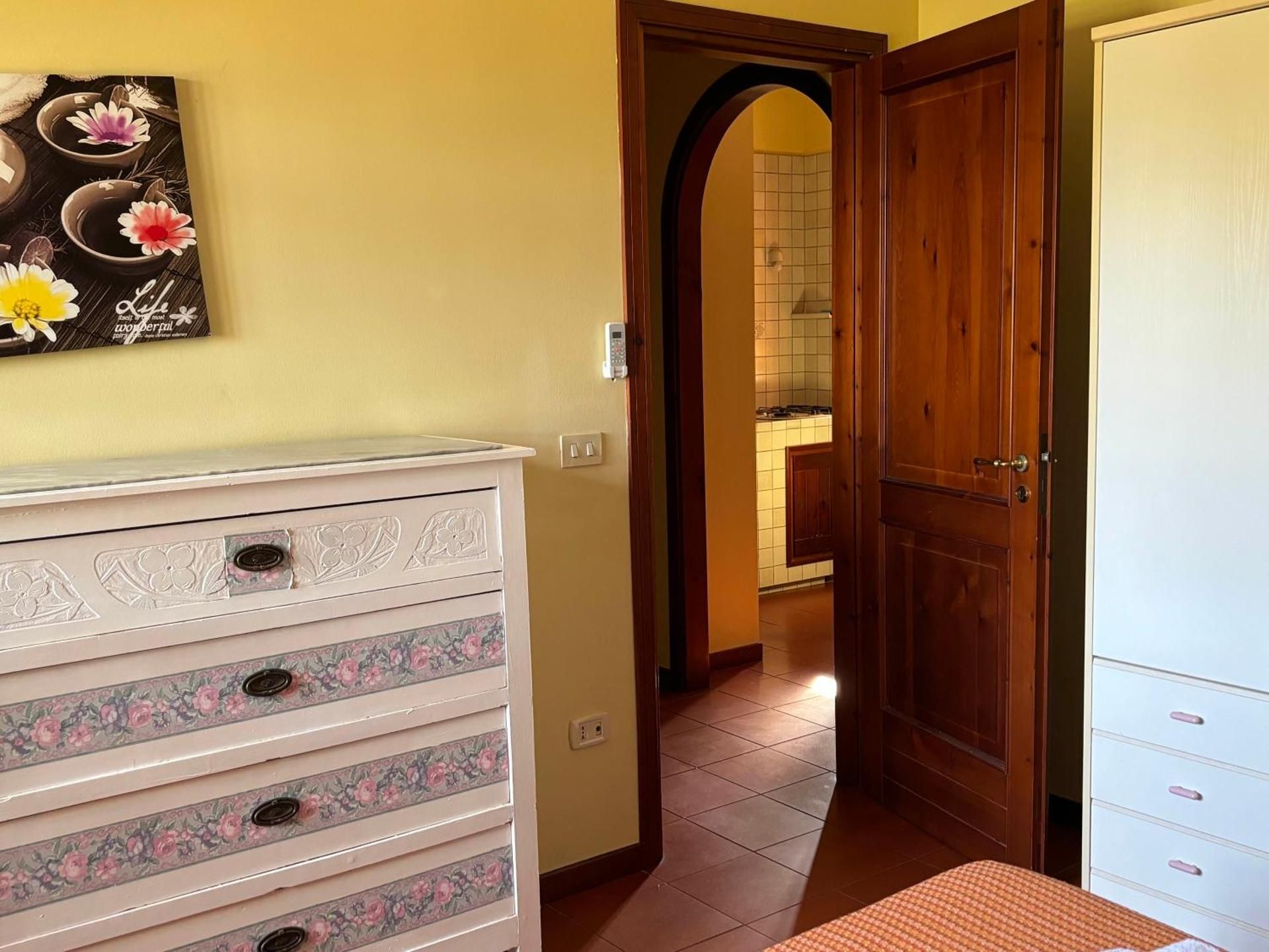 Camera da letto luminosa con comò bianco a motivi floreali, porta in legno e porta ad arco che conduce a un bagno piastrellato.