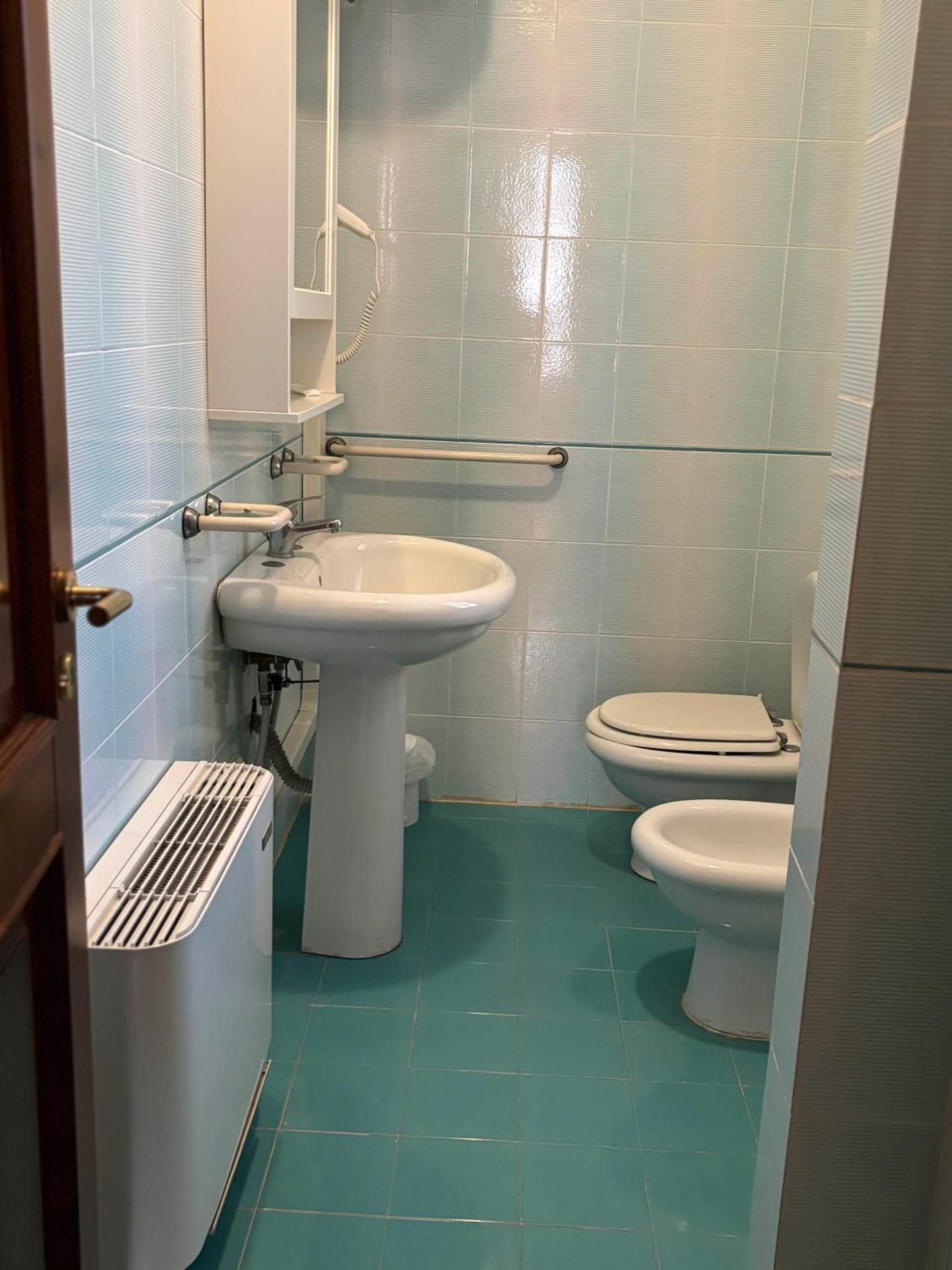 Piccolo bagno accessibile con piastrelle blu, lavabo a colonna, WC e maniglioni di sostegno.