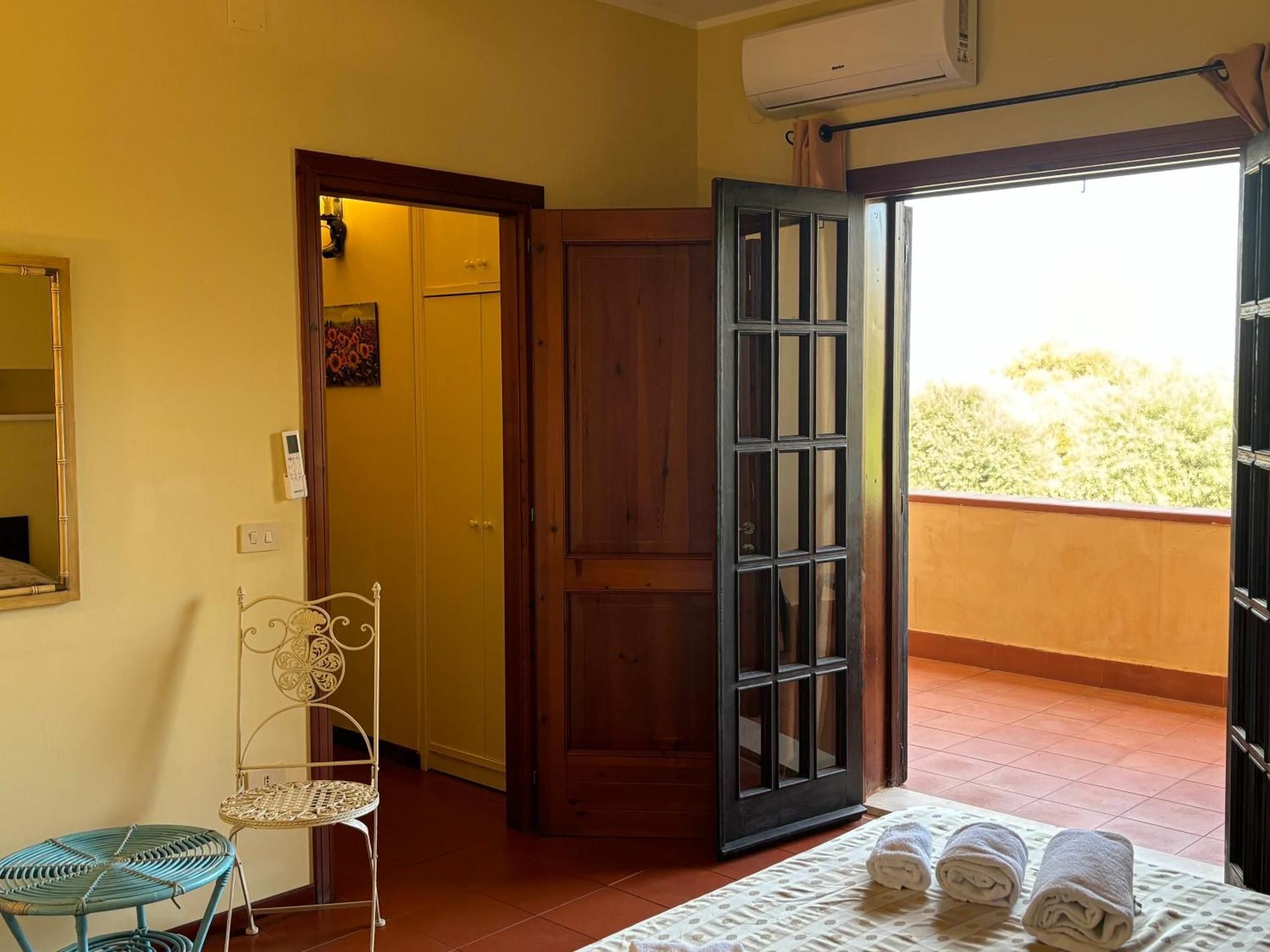 Camera luminosa con porta in legno aperta su un balcone soleggiato e asciugamani piegati sul pavimento.