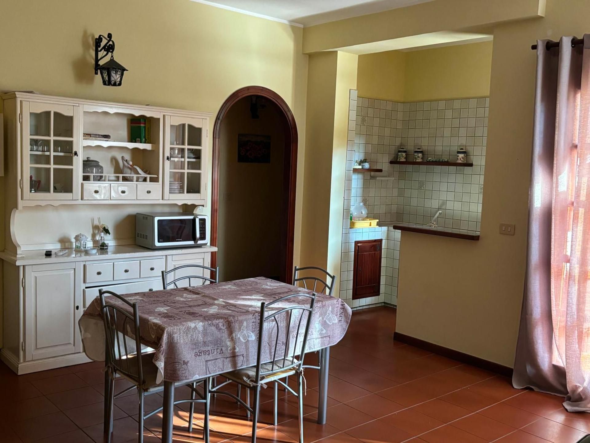Cucina accogliente con mobili bianchi, tavolo da pranzo, arco e tende rosa accanto a una parete piastrellata.