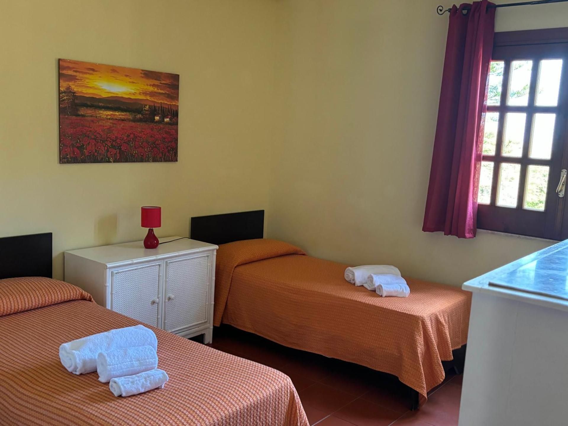 Tre letti singoli in una semplice camera d'albergo con finestra, tenda e quadri alle pareti.