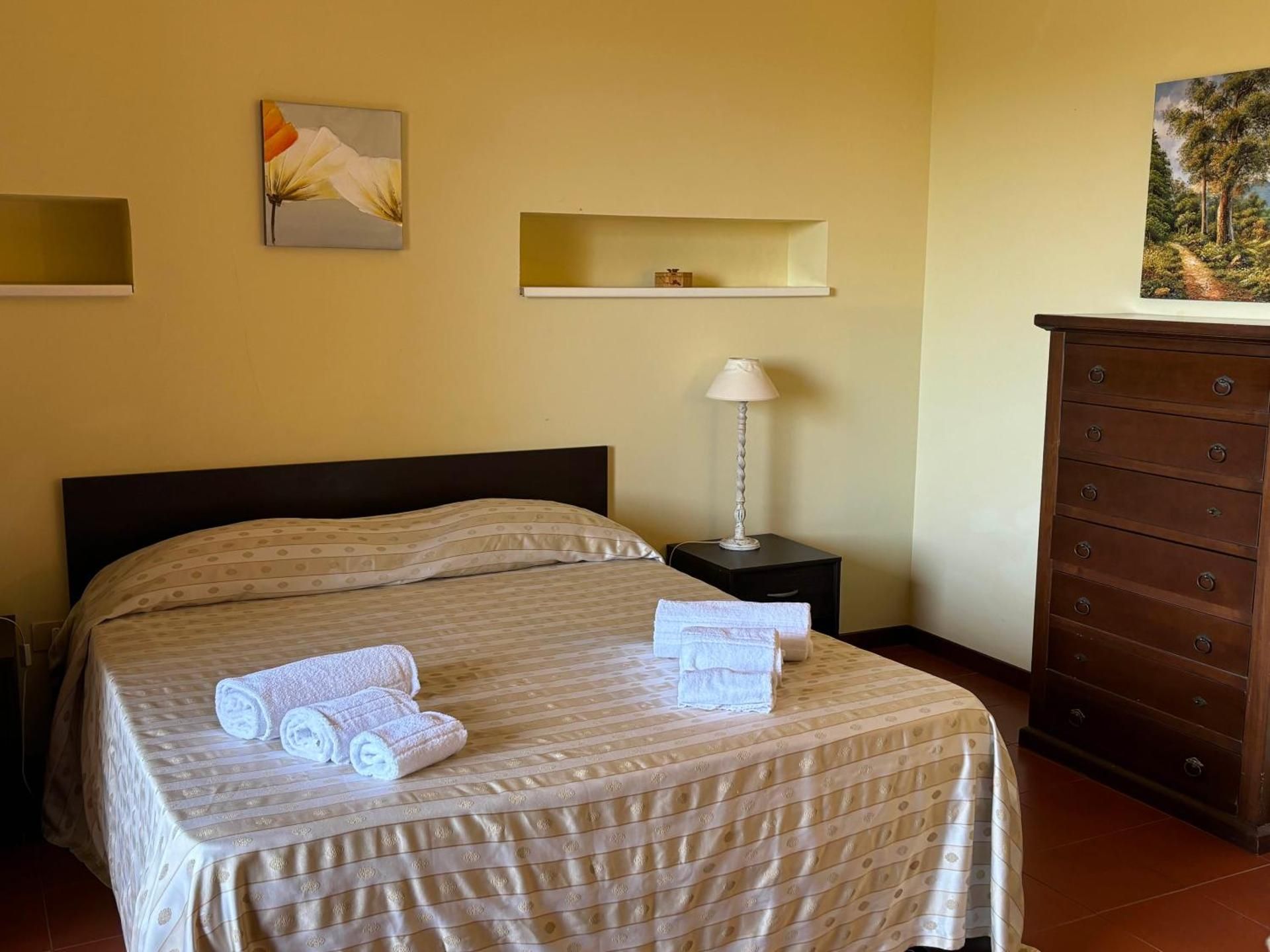 Camera da letto con letto rifatto, asciugamani arrotolati, lampada da comodino, mensola a muro e cassettiera in legno.