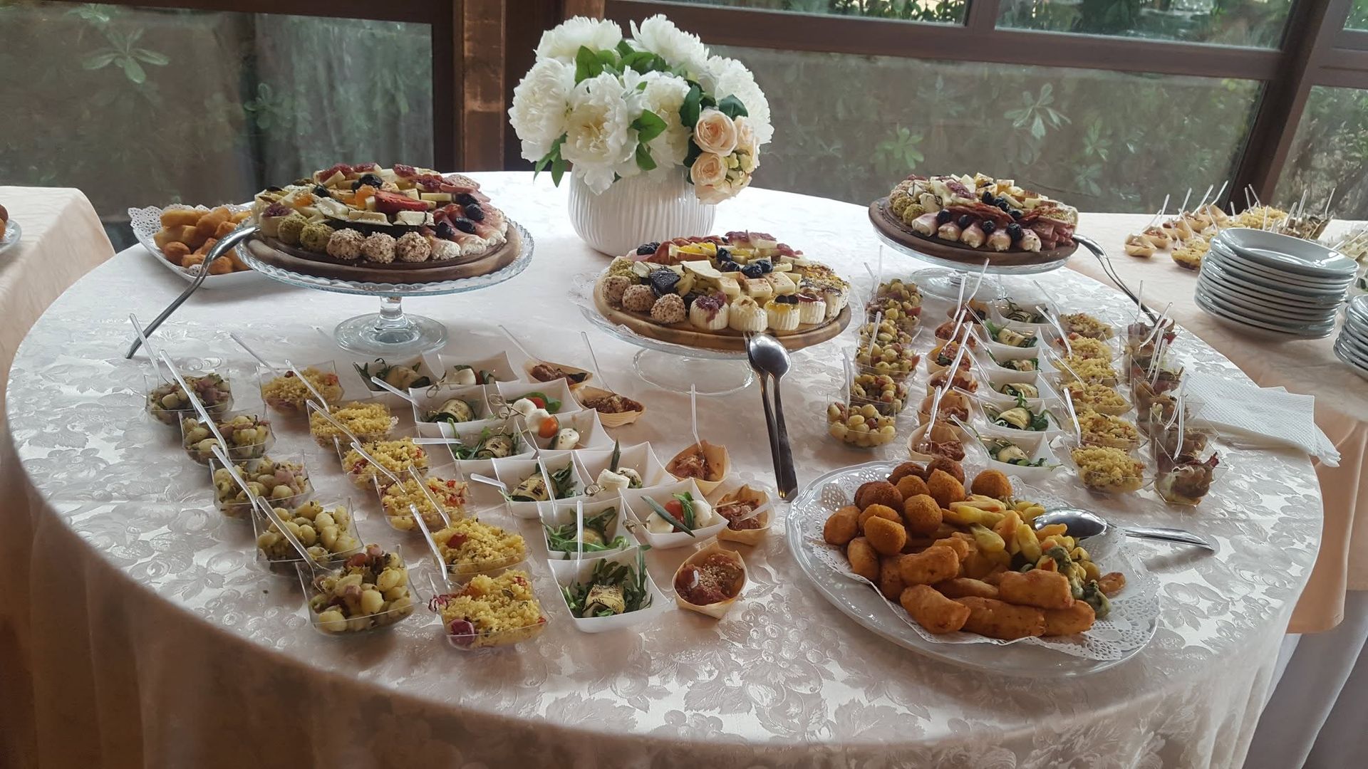 Tavolo rotondo per banchetto con un assortimento di antipasti in piccole tazze e vassoi, al centro del quale è posta una composizione di fiori bianchi.
