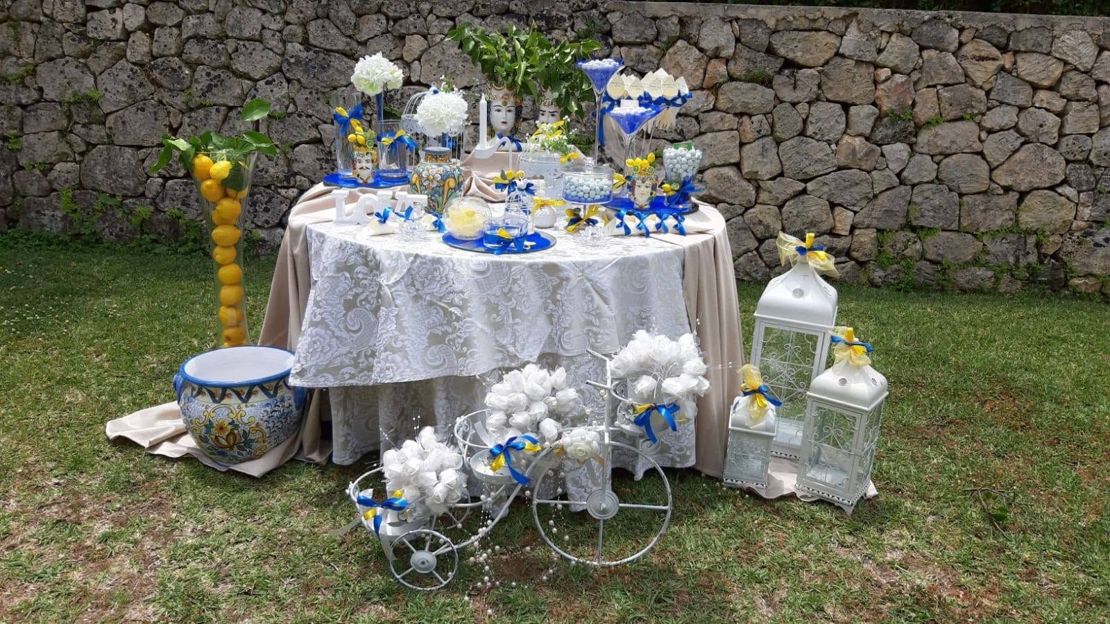 Tavolo da esterno decorato con composizioni floreali blu e bianche, candele e tovaglia di pizzo, addossato a un muro di pietra.