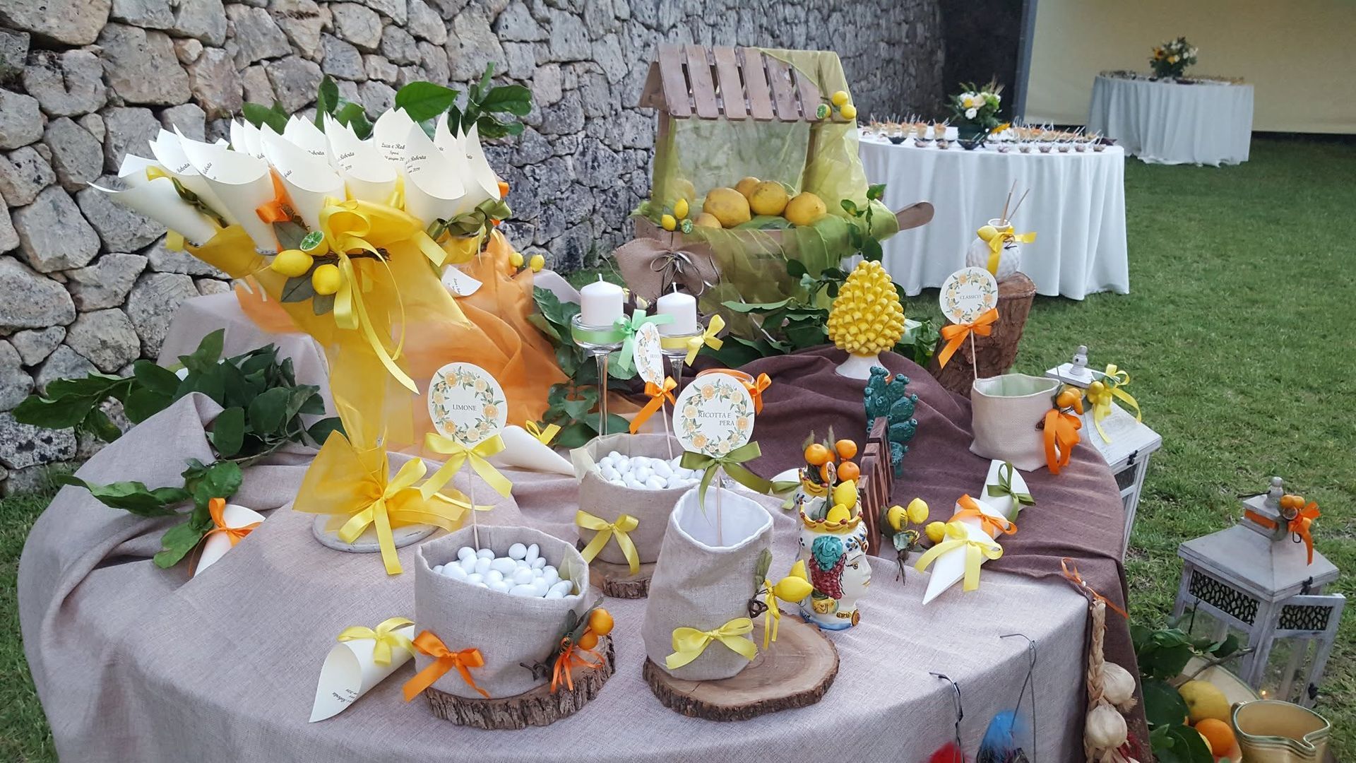 Tavolo all'aperto decorato con centrotavola floreali gialli e bianchi, torte e cesti regalo sull'erba.