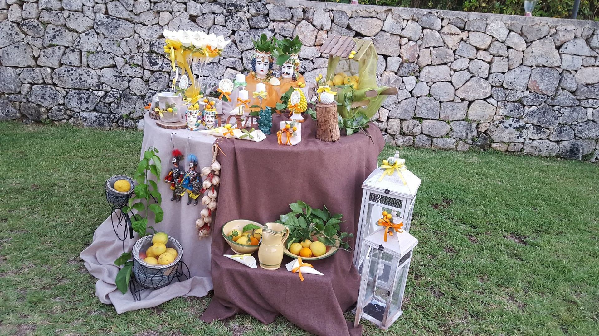 Allestimento esterno con allestimento di prodotti del raccolto su un tavolo ricoperto da una tovaglia marrone, con zucche, zucchette ornamentali, frutta, fiori e lanterne, accanto a un muro di pietra.